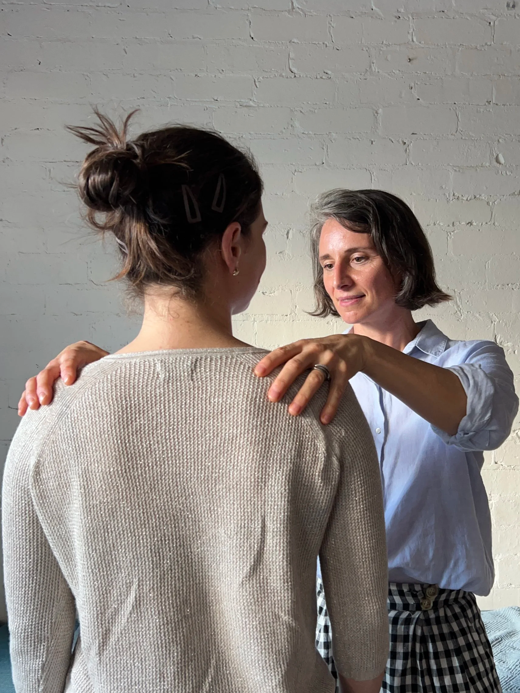 Two women are standing close together against a white brick wall. One woman, with gray hair and a white shirt, is facing the other woman, who has brown hair in a bun, wearing a beige sweater and has the back to the camera. The woman with gray hair has her hand on the other woman's shoulder.