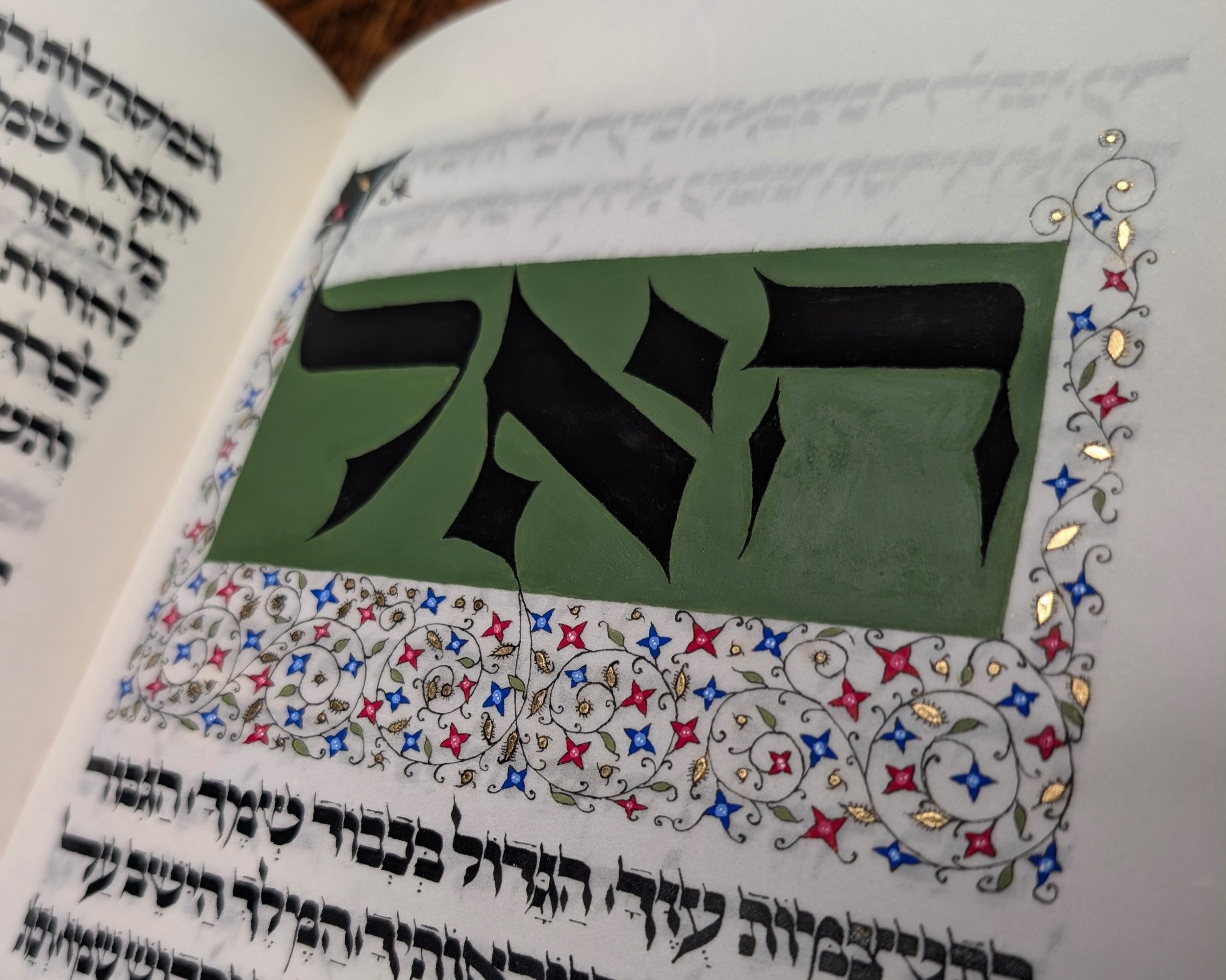 A colorful custom illuminated Haggadah (haggada) Hebrew manuscript page in the medieval Ashkenazic style with black Hebrew letters, decorative floral border with red, blue, and gold accents, and an olive green background behind the letters.