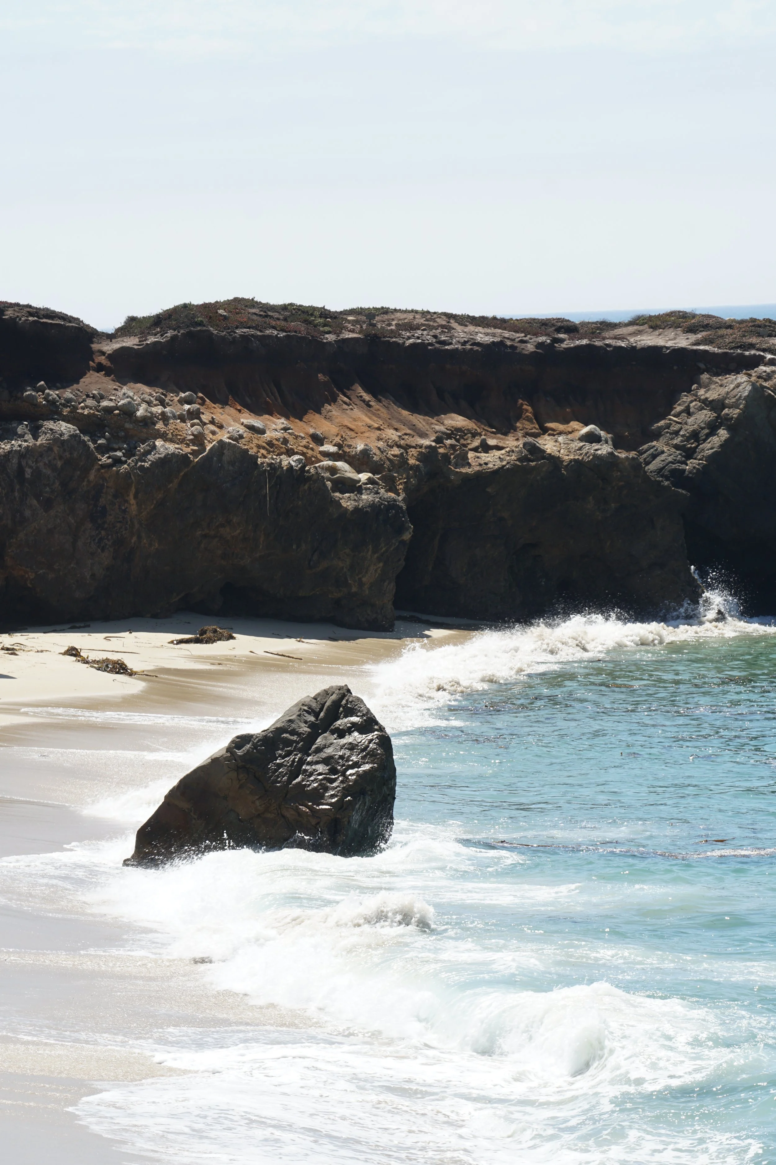 Coastal scene with large rocks on sandy beach and cliffs in the background under a clear sky.