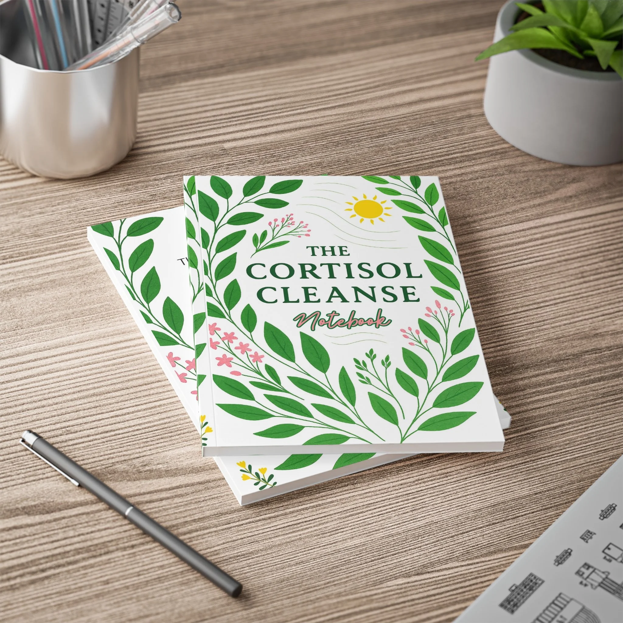 Close-up of a notebook titled "The Cortisol Cleanse Notebook" on a wooden desk, surrounded by a pen, a potted plant, and a container holding pens and markers.
