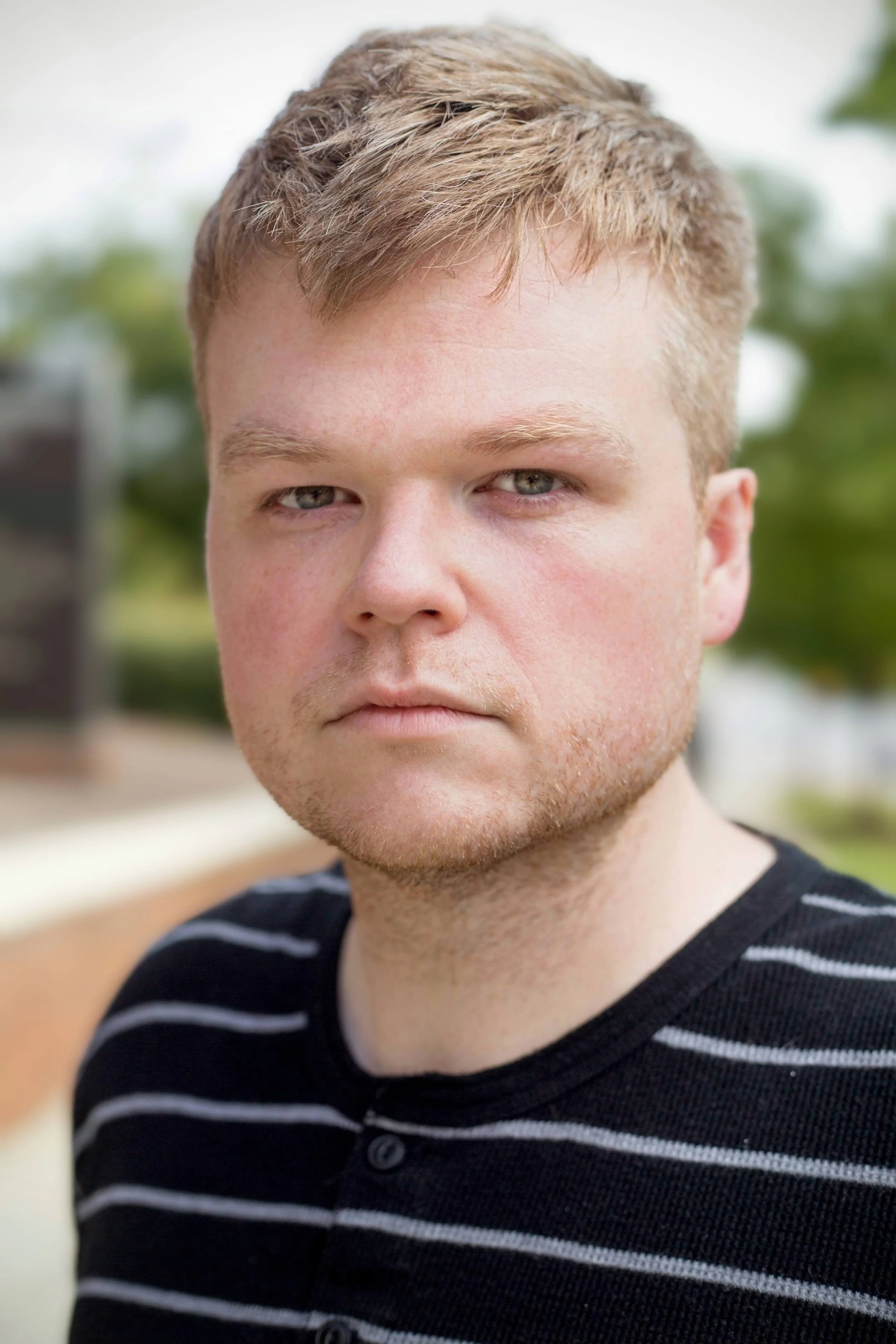 Portrait of artist Benjamin Jones wearing a black striped shirt against a softly blurred outdoor background.