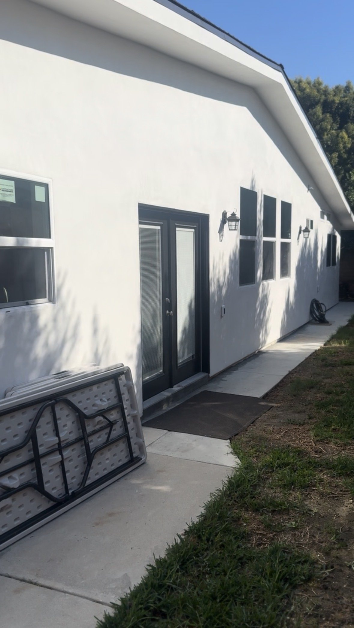 Back exterior of a white house with a black door, several windows, wall-mounted lanterns, and outdoor furniture on the concrete patio.