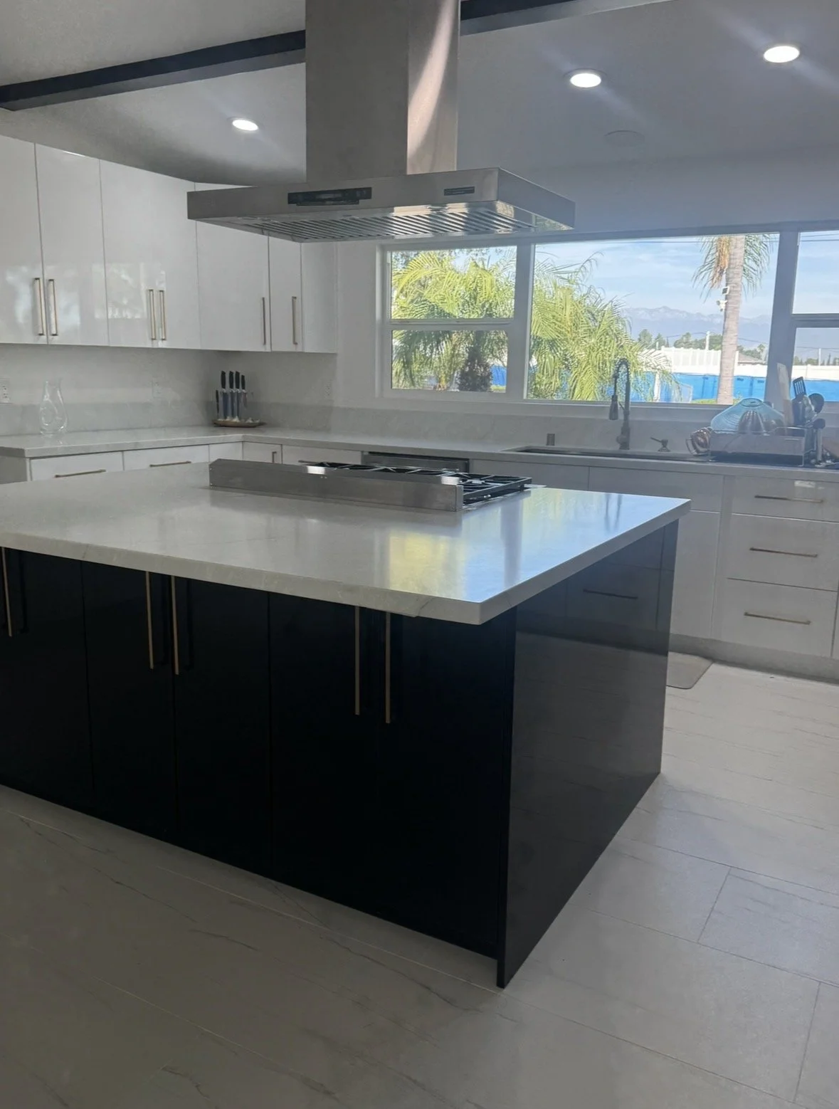 Modern kitchen with white countertops, a black island with gold handles, stainless steel stove hood, and large window showing trees and a blue fence outside.
