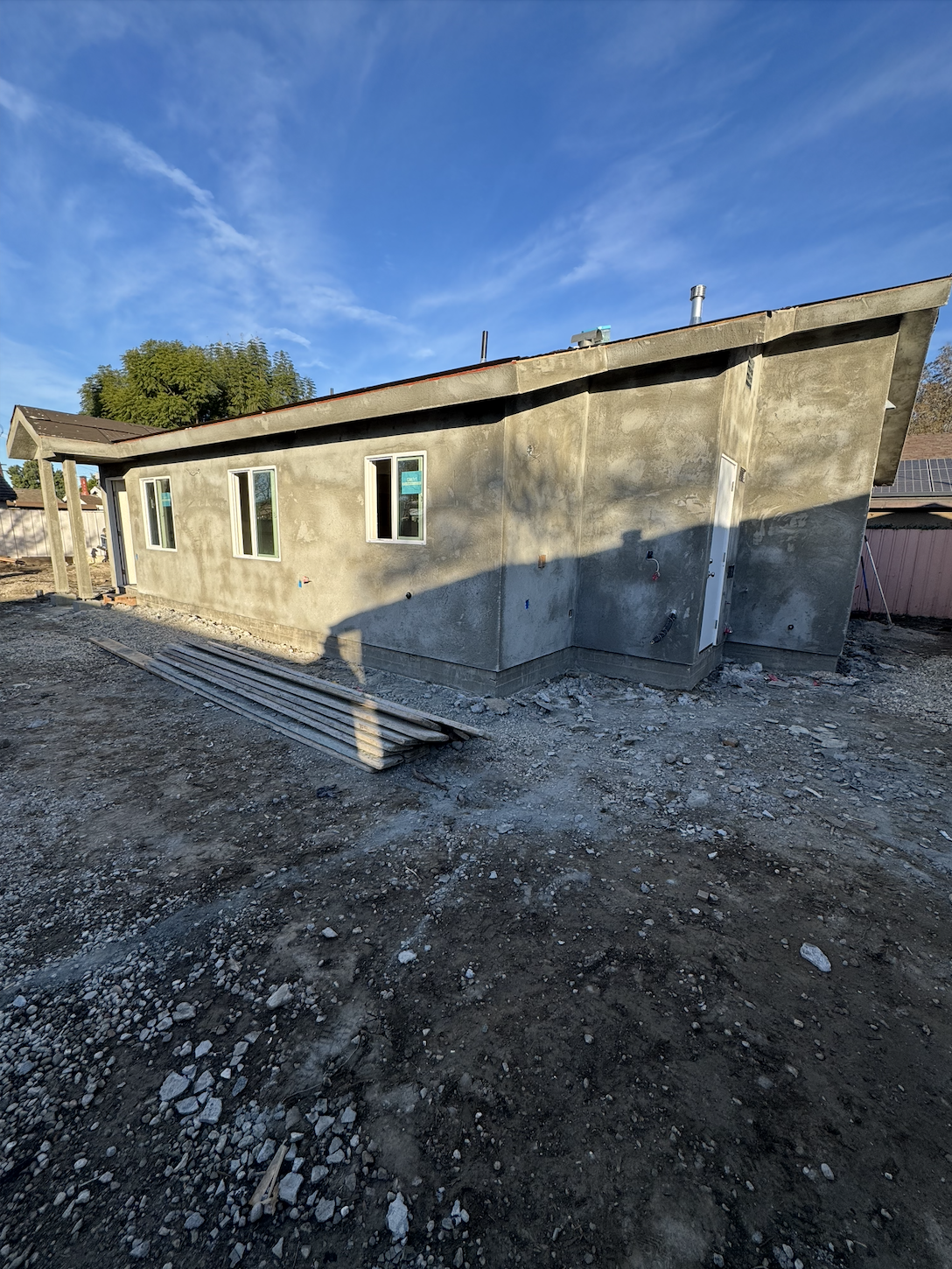 Construction site of a single-story house with unfinished exterior walls, four windows, a door, and a sloped roof, surrounded by dirt and construction materials.
