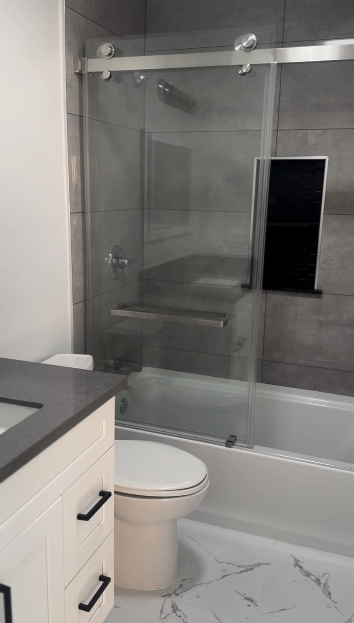 A modern bathroom with a white toilet, a dark gray countertop with a sink, a glass enclosed shower with gray tiles, a bathtub, and a black wall shelf.