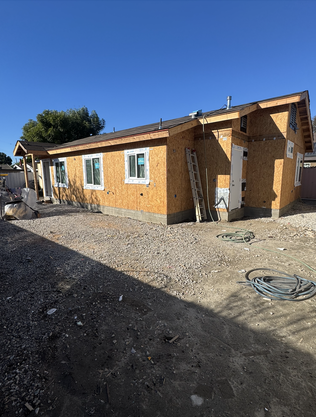 A house under construction with wooden exterior walls, multiple windows, and a sloped roof. Construction materials and tools are visible around the site, with a clear blue sky overhead.
