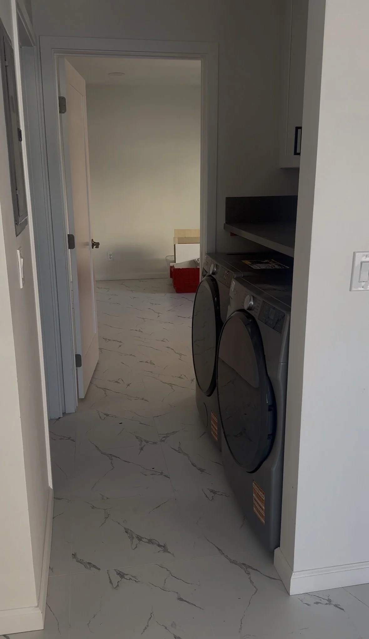 Laundry room with a front-loading washing machine and dryer, white marble floor, and a doorway leading to a small room with a bed frame and red storage bin.