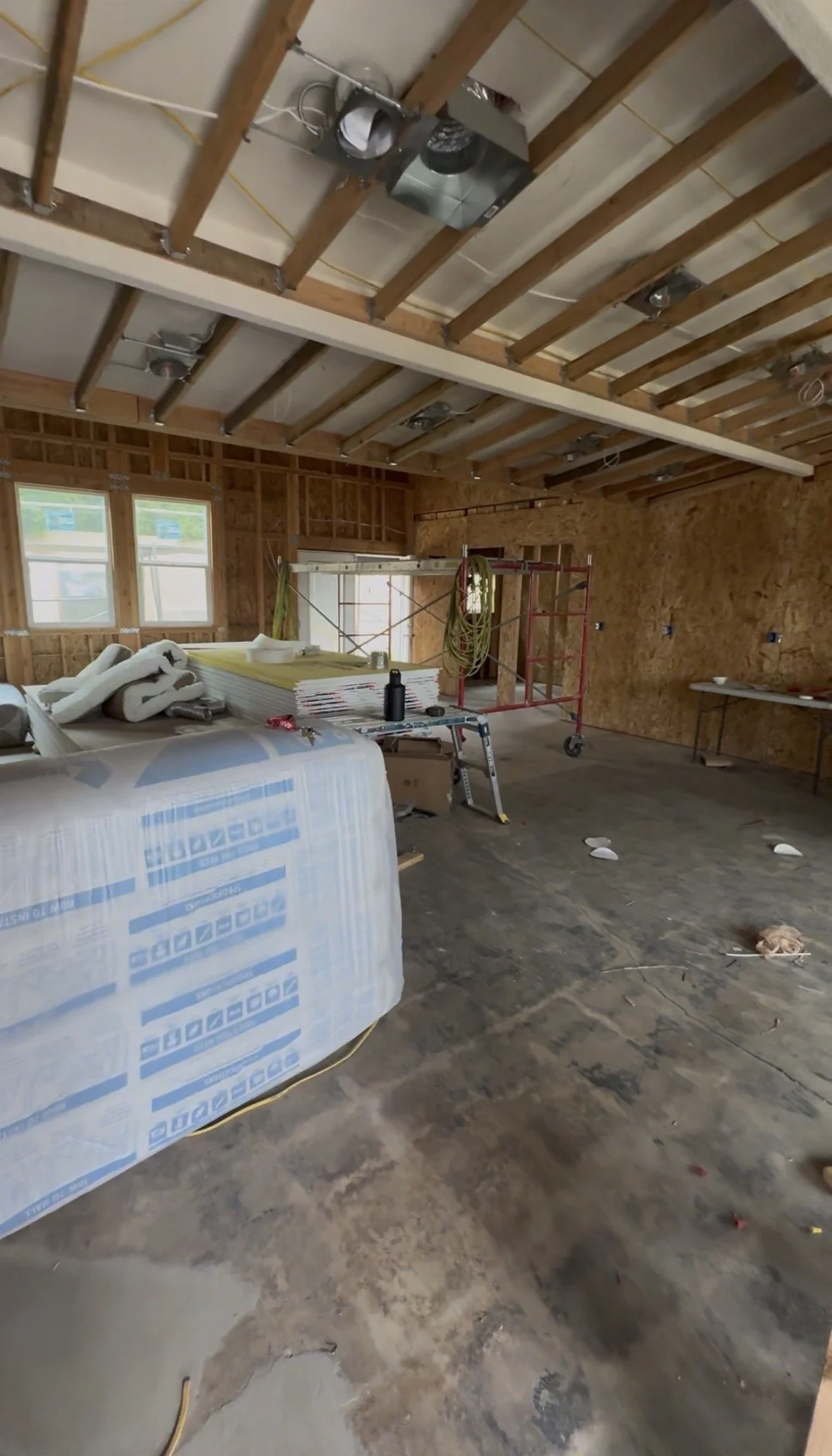 Construction site of a house interior with unfinished walls and ceiling, scaffolding, construction materials, and tools scattered on the floor.