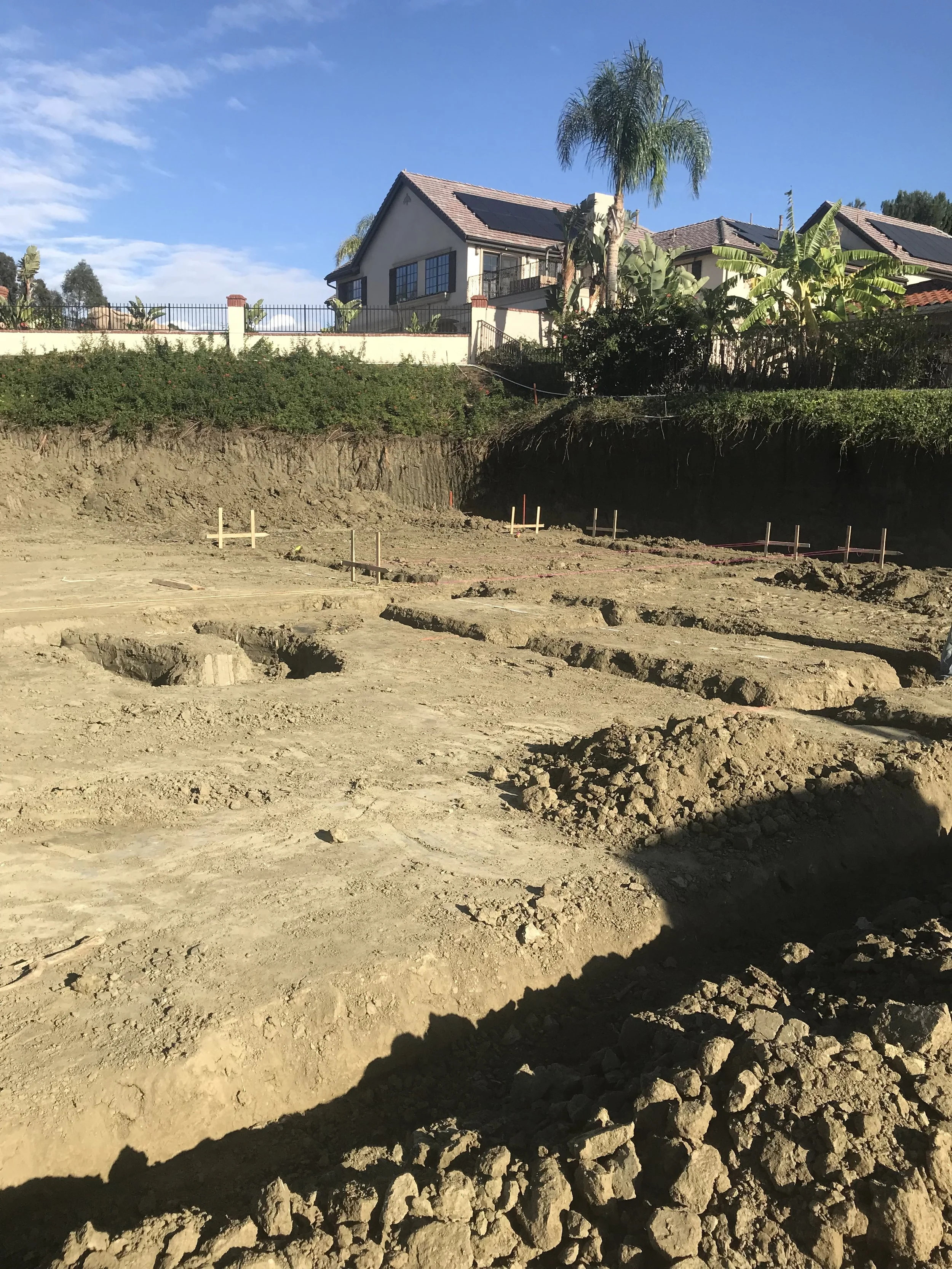Construction site with excavated dirt and groundwork, residential houses with palm trees in the background, clear blue sky.