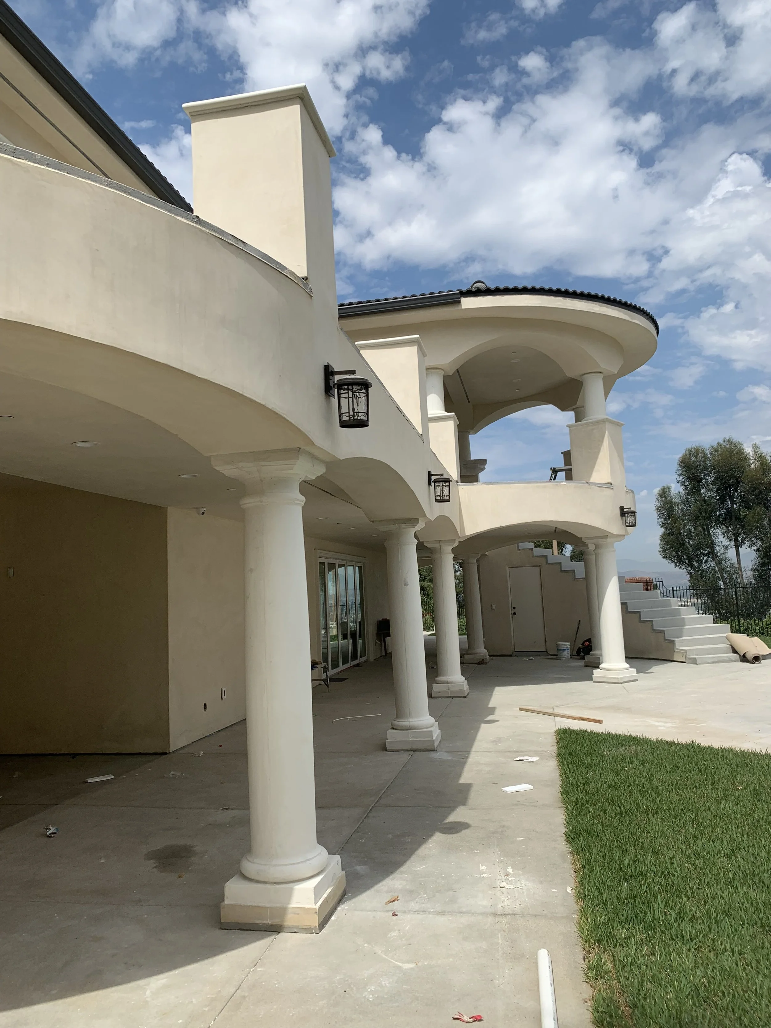 A multi-story house under construction with white stucco exterior, rounded balconies, and decorative columns. The patio area is partially finished, with some tools and materials scattered around. There is a grassy patch and stairs leading to the uppe