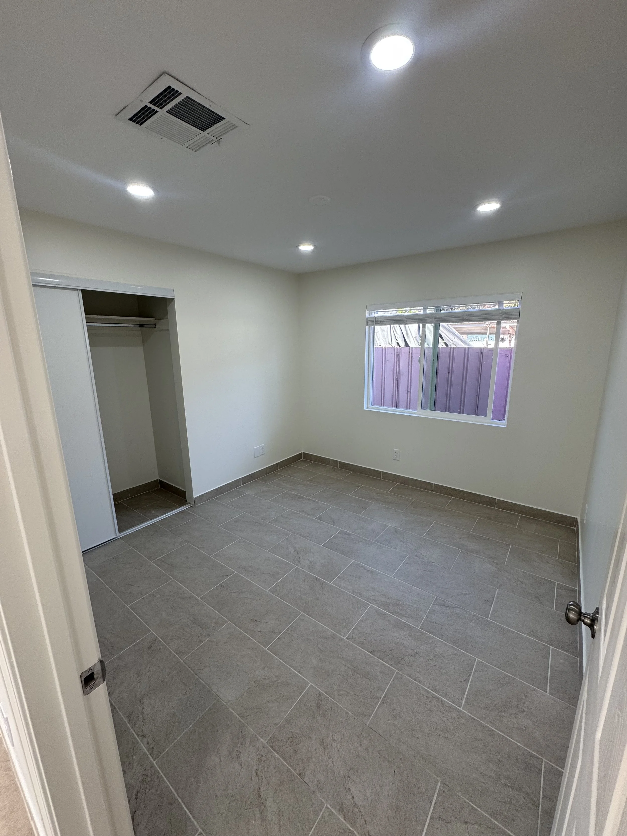 Empty bedroom with beige tiled floor, white walls, a window with blinds, a closet with sliding door, and ceiling lights.