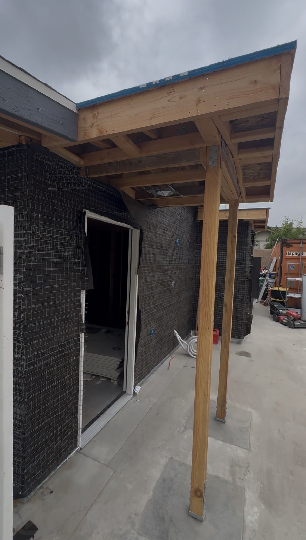 Side view of a house exterior under construction with a wooden porch roof, black waterproofing, and construction tools and materials nearby.