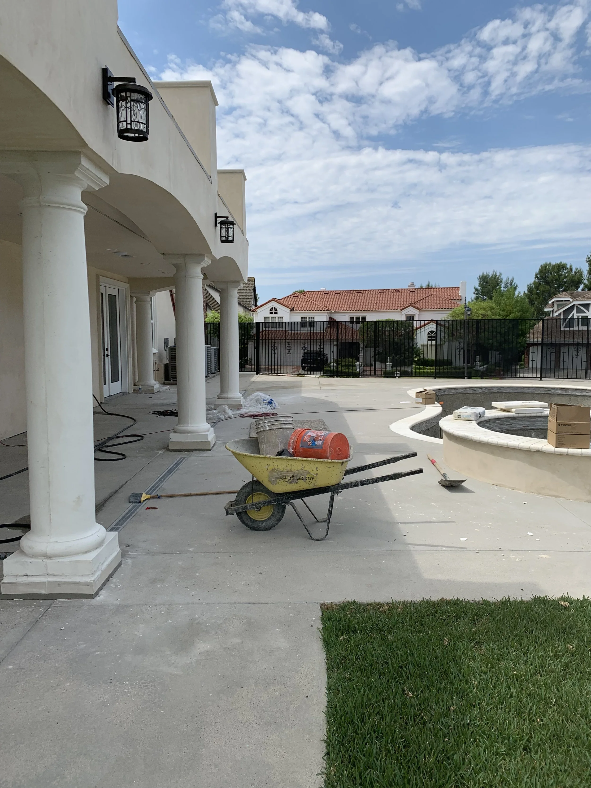 Construction site in backyard with wheelbarrow carrying buckets and a gas can, pool area under construction, and neighboring houses with red tile roofs.