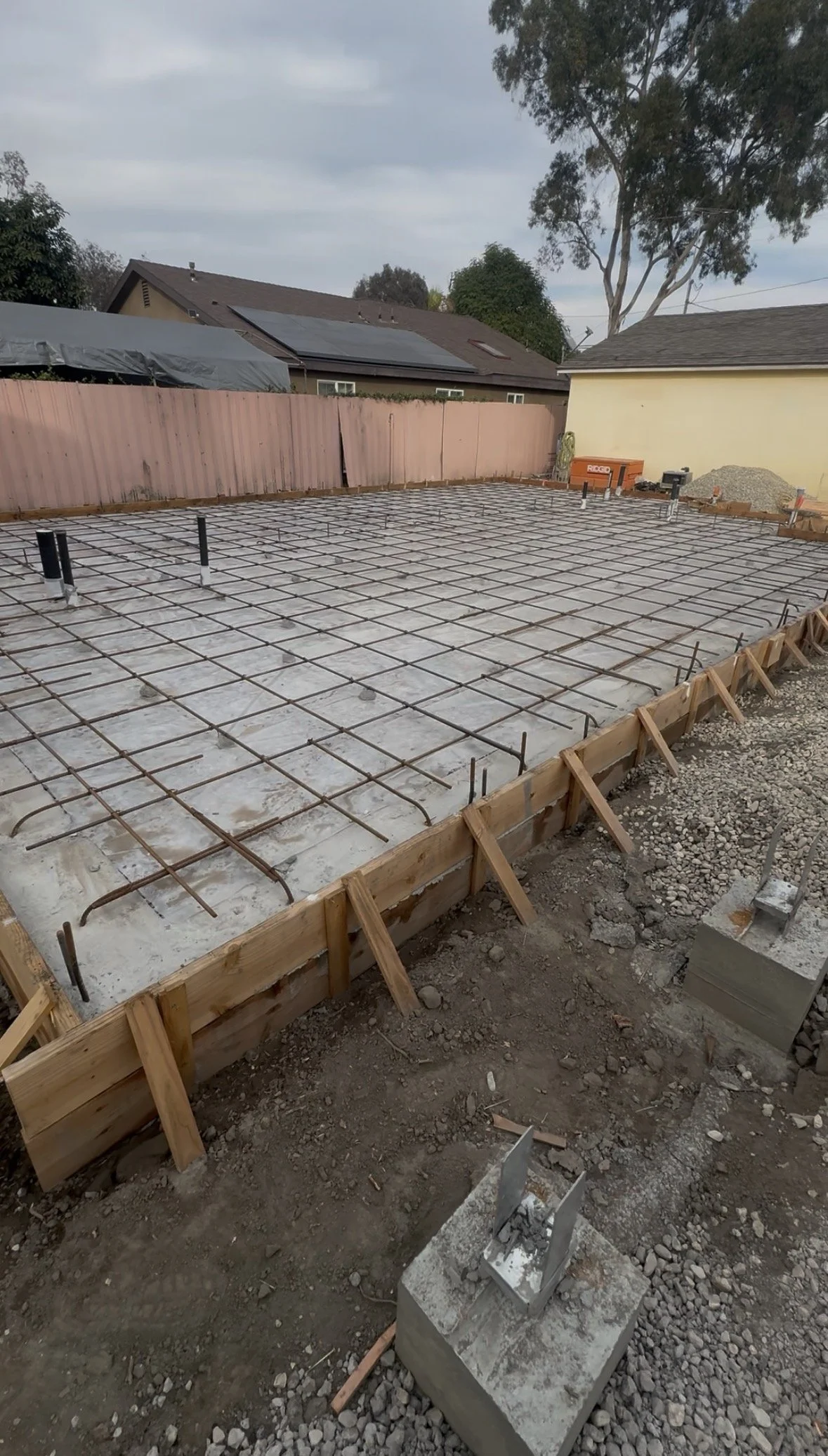 Construction site with concrete slab and rebar reinforcement for building foundation, surrounded by wooden framing, with neighboring houses and trees in the background.