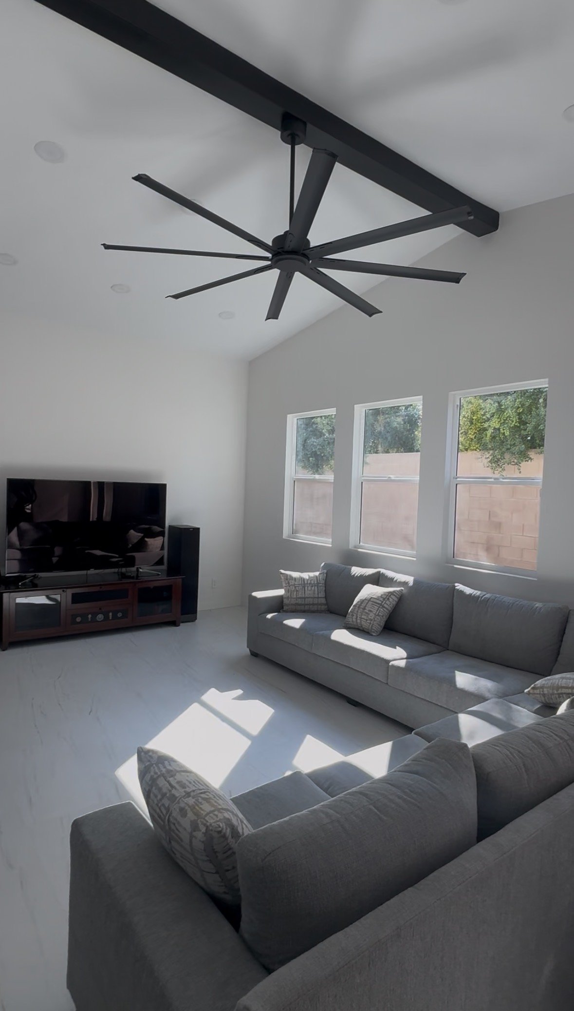 Living room with a ceiling fan, gray sofas, a flat-screen TV on a stand, and three windows allowing sunlight to enter.