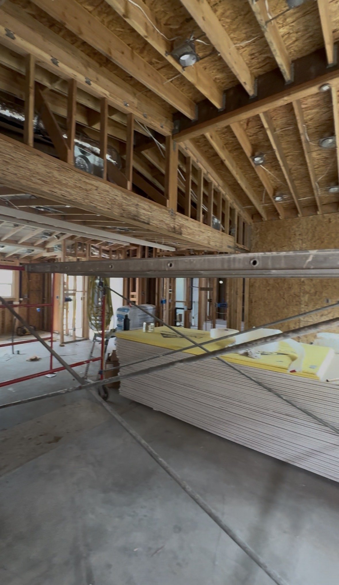 Interior view of a construction site showing exposed wooden framing, plywood walls, a stack of drywall sheets, scaffolding, and construction debris.