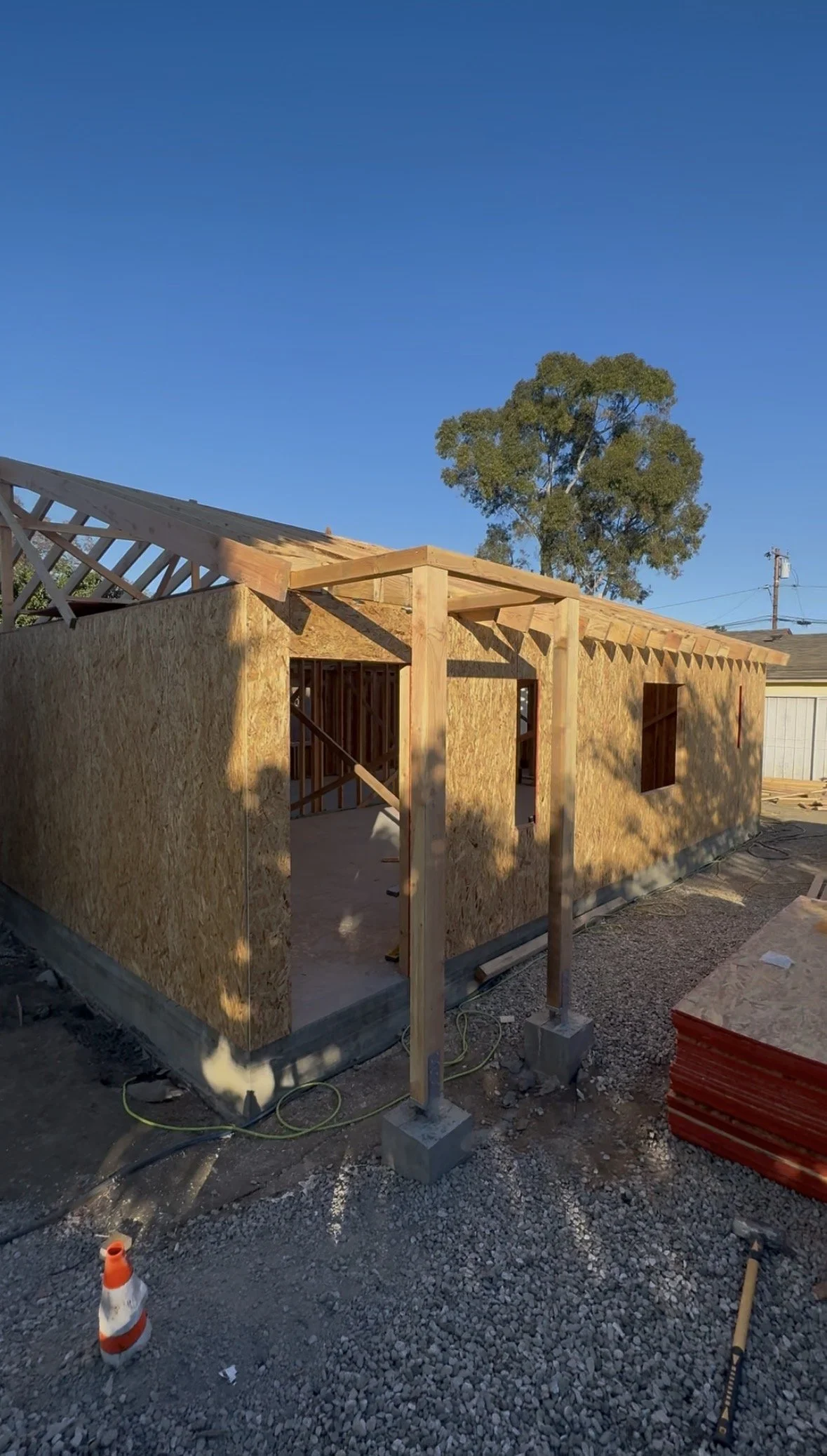 Wooden framework and siding of a house under construction with a clear blue sky and a tree in the background.