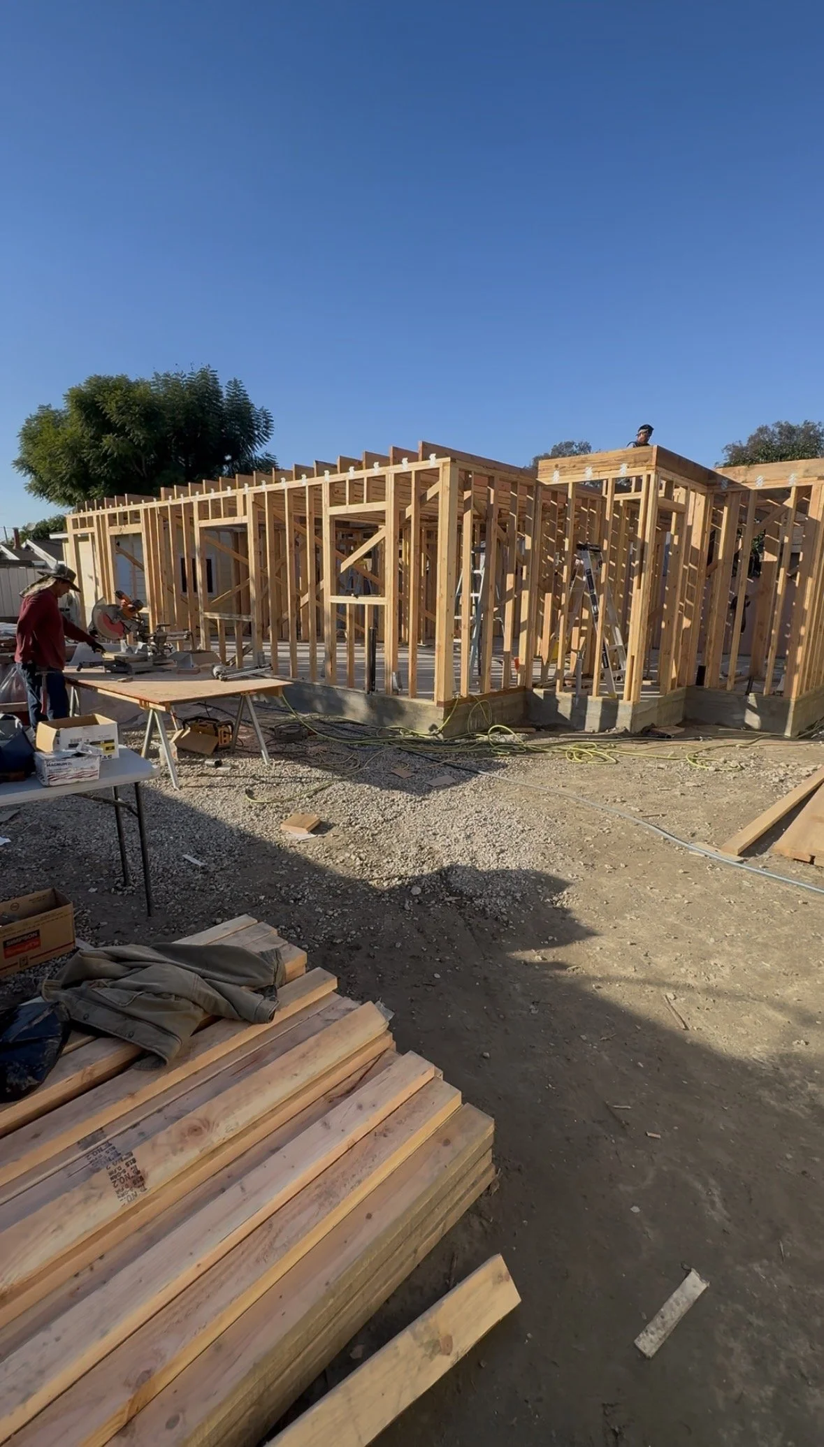 Construction site with wooden framing for a building, workers, tools, and construction materials on the ground under a clear blue sky.