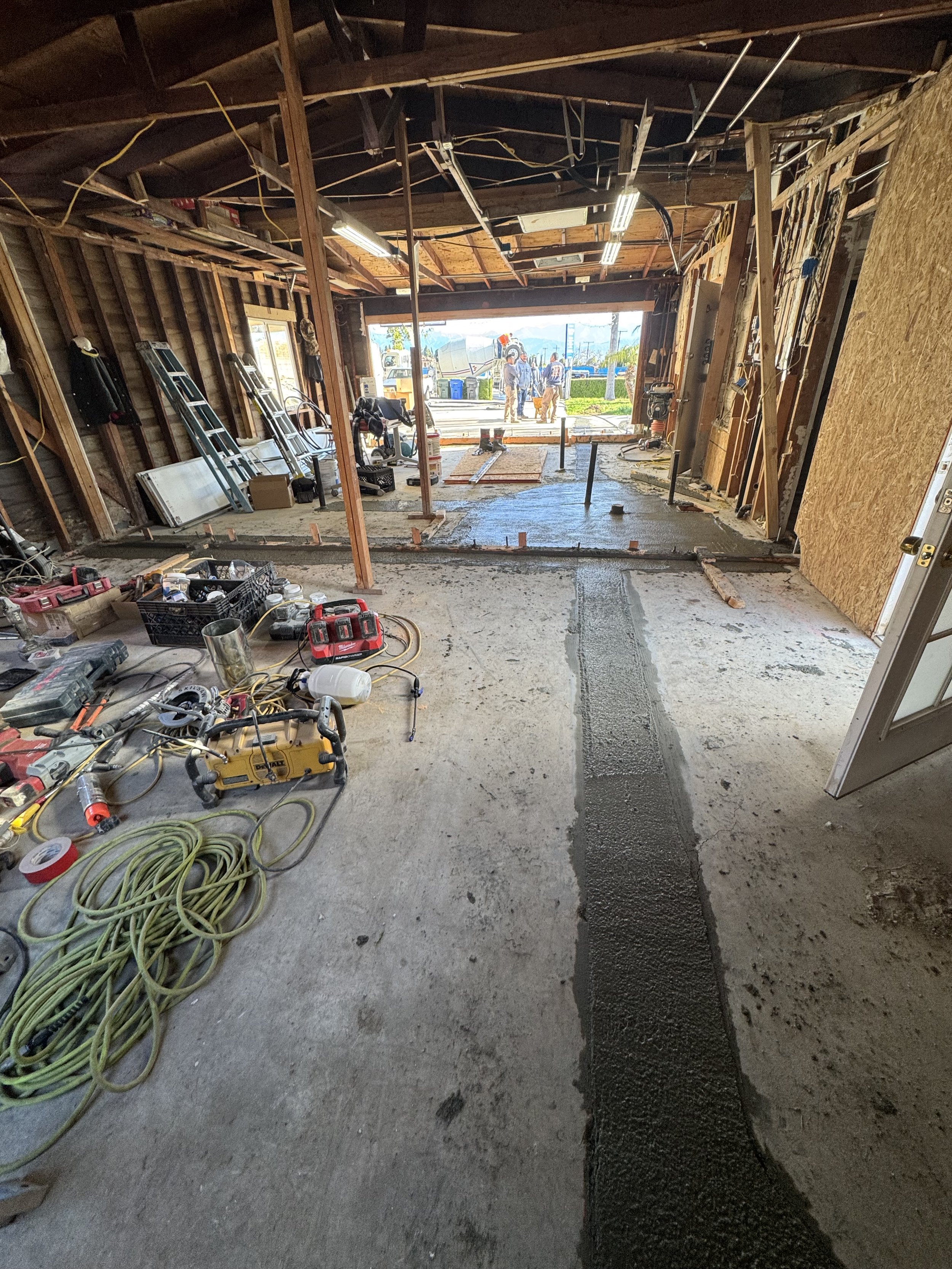 Garage under construction with tools, equipment, and building materials scattered around. A wet concrete path is being poured in the center, with an open garage door revealing a street scene and workers outside.