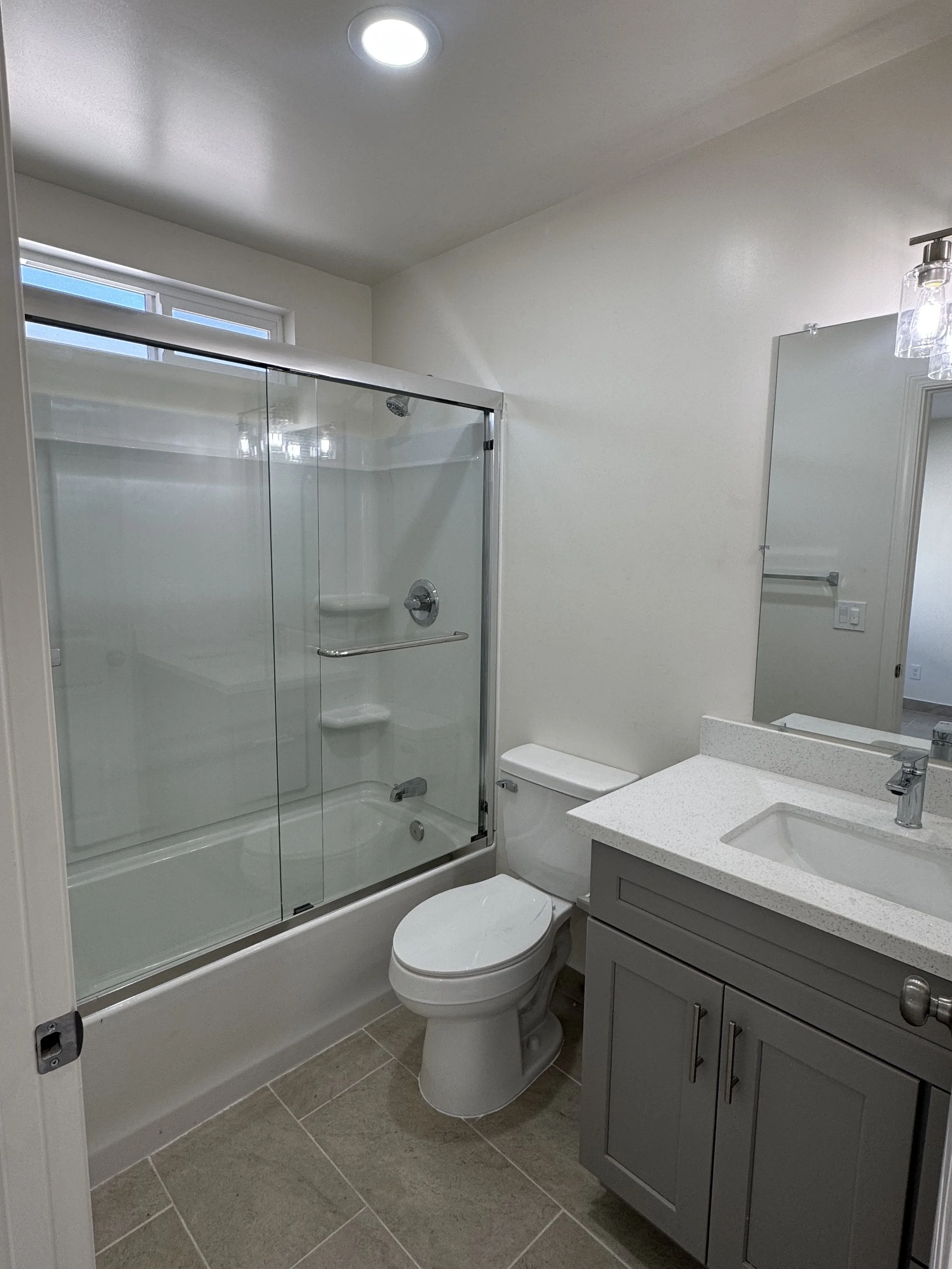 A modern bathroom with a bathtub and shower combination enclosed with glass doors, a white toilet, and a grey vanity with a white countertop and sink. A mirror and wall-mounted light fixture are above the vanity, and a window is above the shower, all