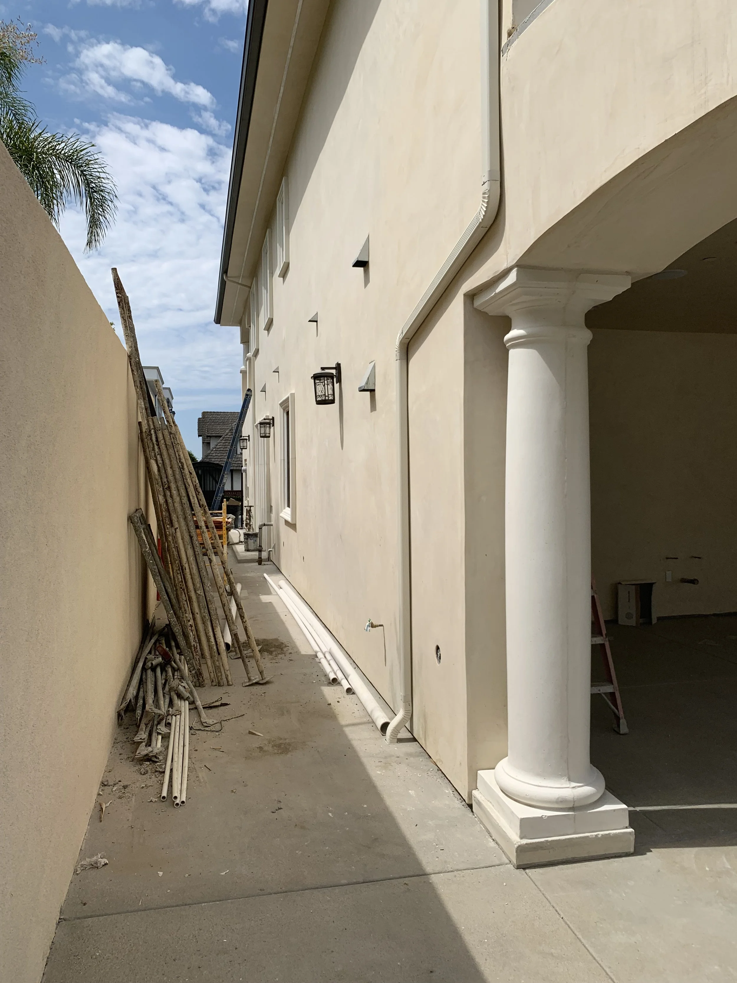 Side view of a beige stucco house under construction with a large white column and shadows cast on the sidewalk, construction materials leaning against the wall, and a partly cloudy sky in the background.