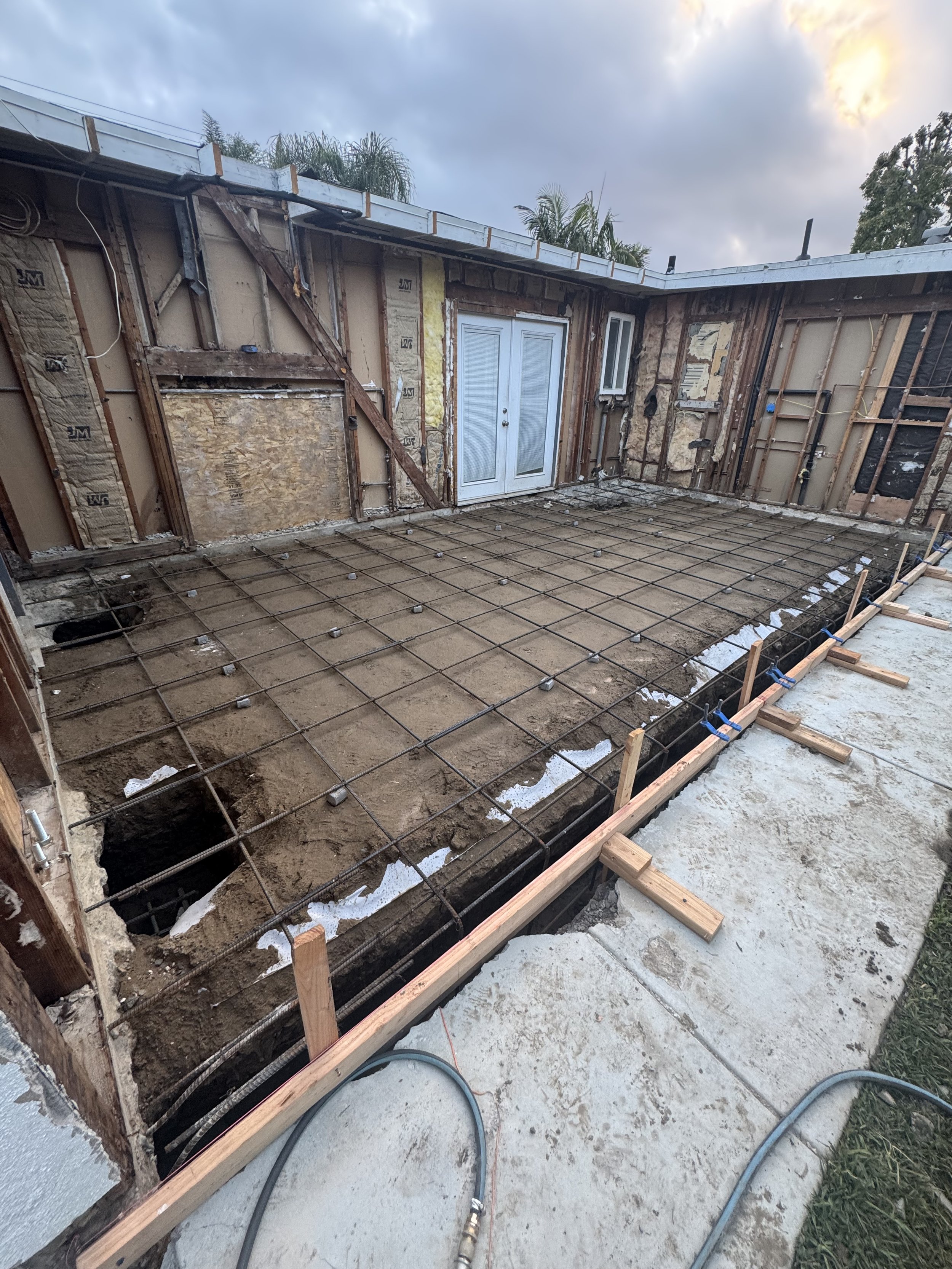 Construction site of a deck with rebar framework for concrete pouring, surrounded by wooden forms, in a backyard with house walls and trees in the background.