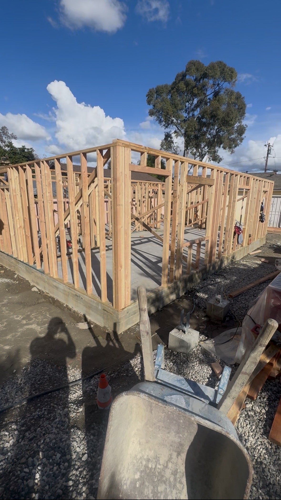 Construction site with wooden framing for a house on a concrete foundation, under a partly cloudy sky, with scaffolding and construction materials around.