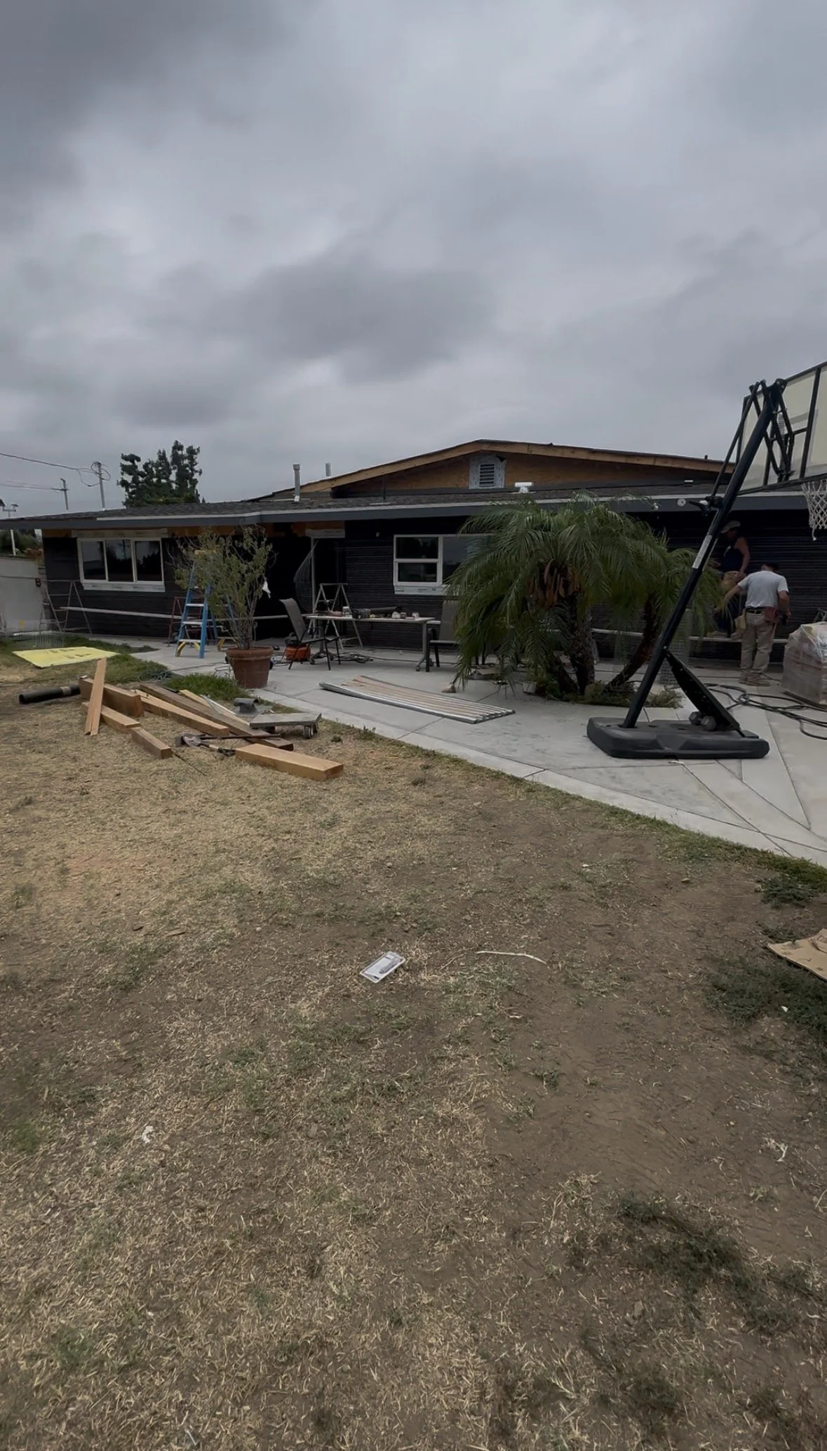 A backyard under construction with a partially completed house, construction workers, scattered wood, potted plant, and a basketball hoop, under cloudy skies.