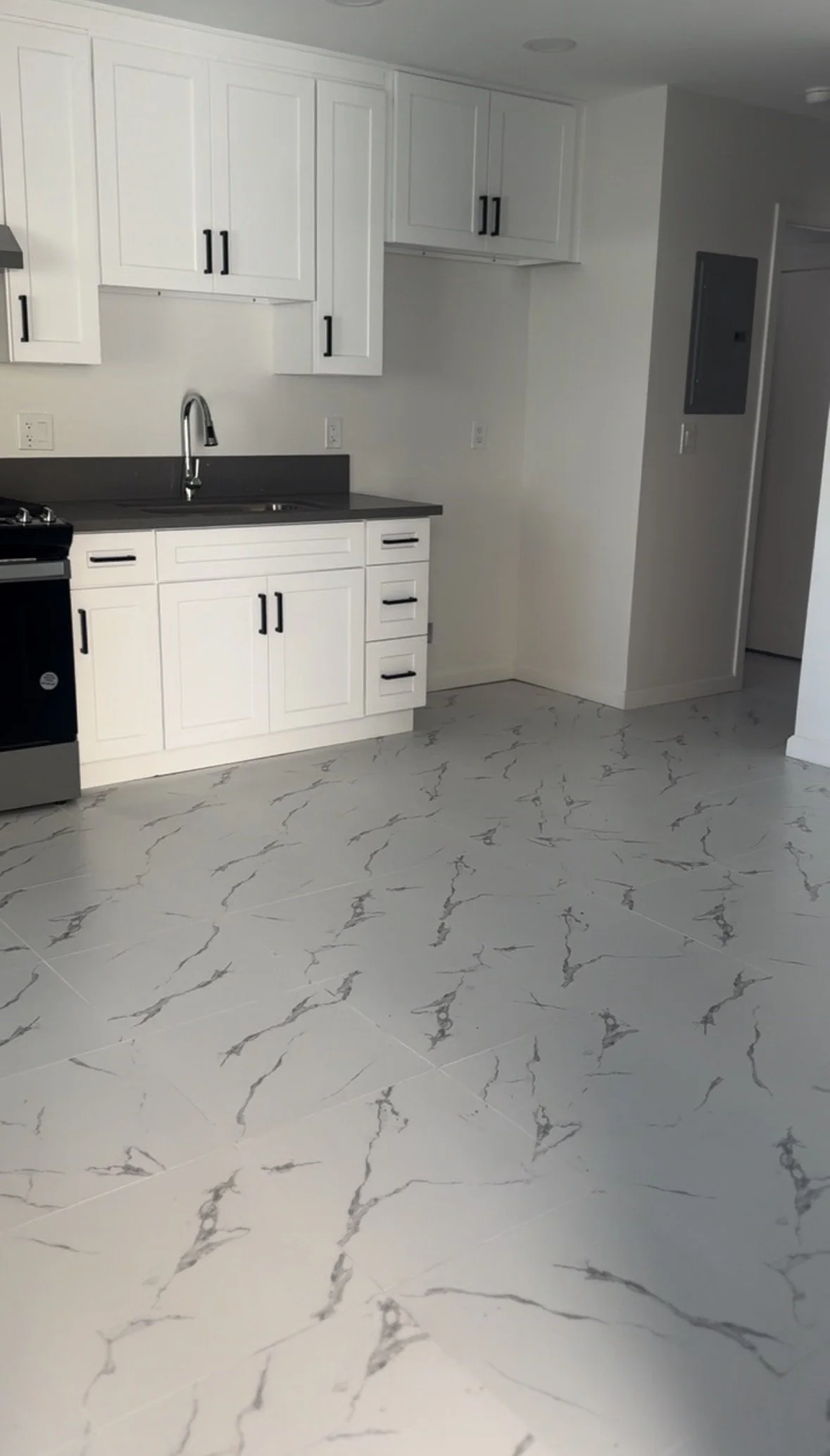 Modern kitchen with white cabinets, black hardware, and a dark countertop. Light-colored marble-like floor tiles with gray veining. No visible appliances or decorations.