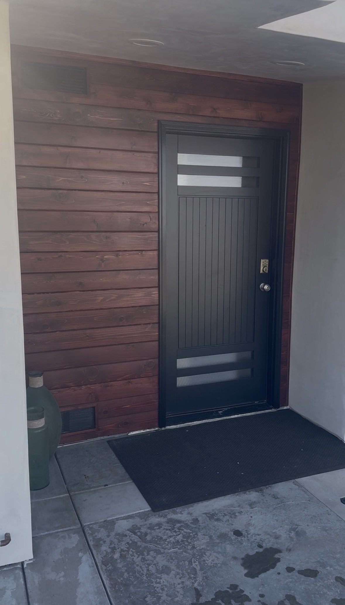 Black security door with horizontal glass panels and a round doorknob, installed on a wooden wall with horizontal planks. A black mat on concrete floor in front of the door and two large vases to the left.