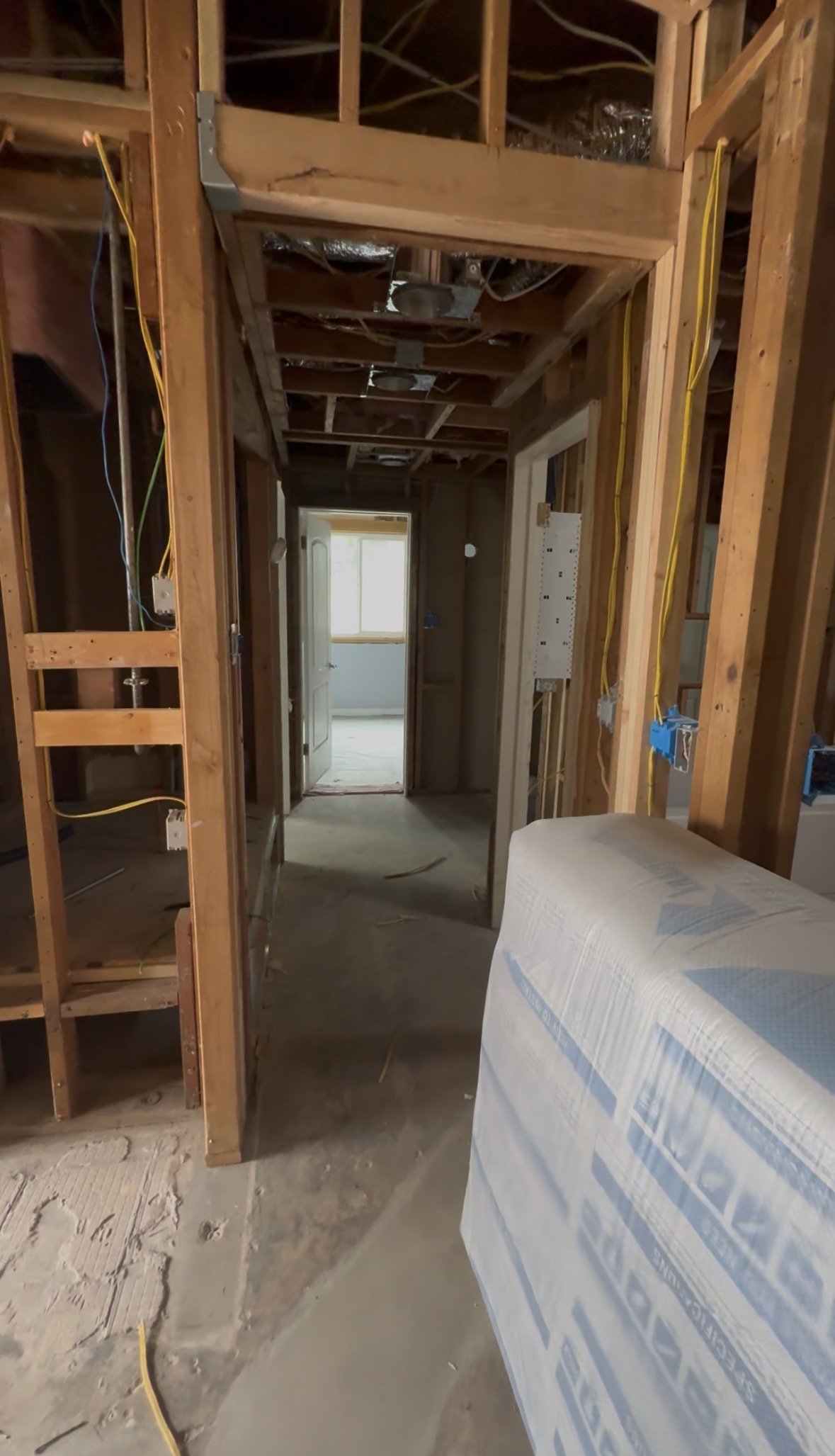 An unfinished interior of a house under construction, showing exposed wooden framing, electrical wiring, and a hallway leading to a room with a closed window.