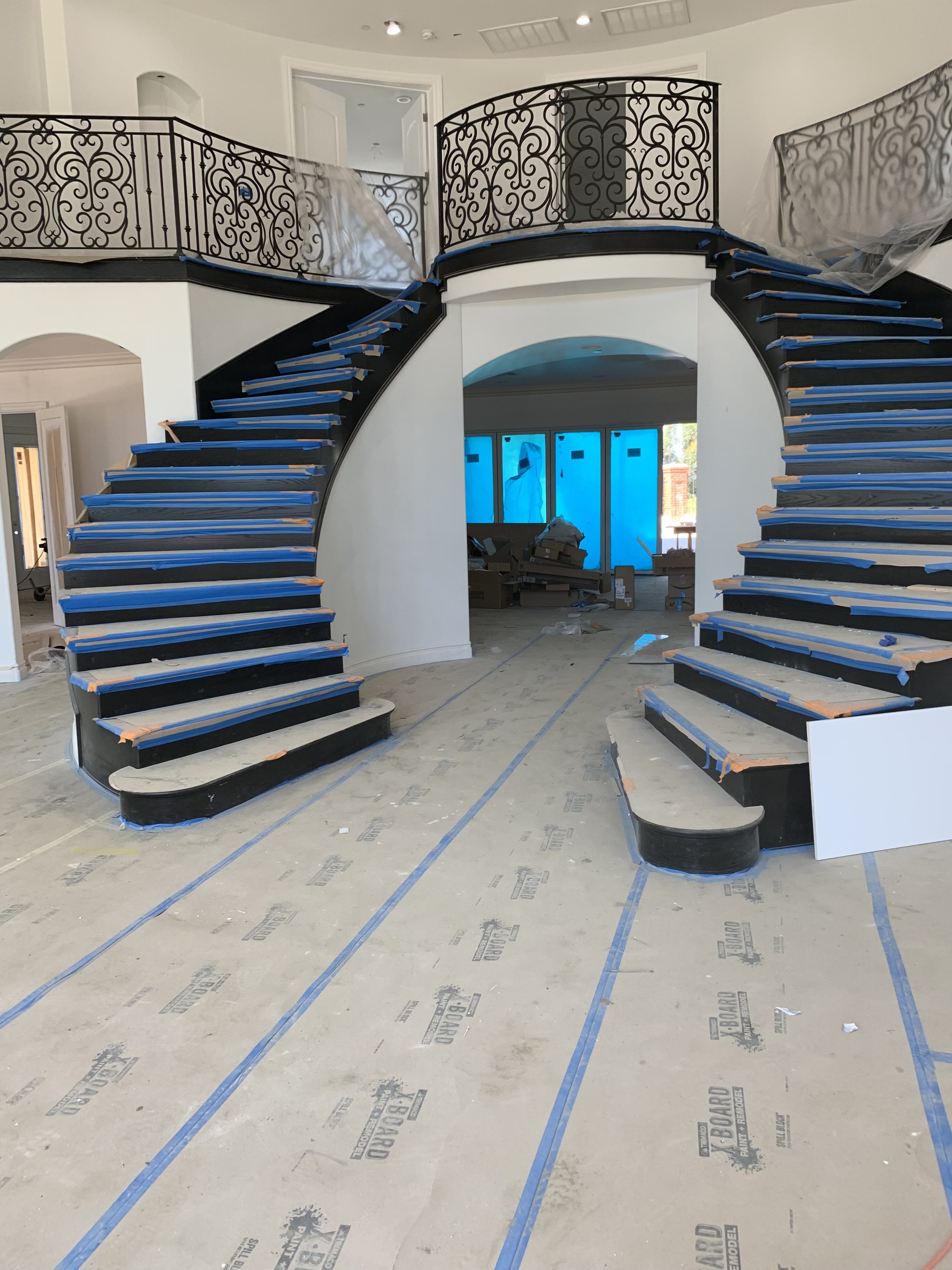Interior of a house under renovation with a curved staircase being painted black, covered with blue painter's tape, and protected with plastic sheeting, with construction materials and tools in the background.
