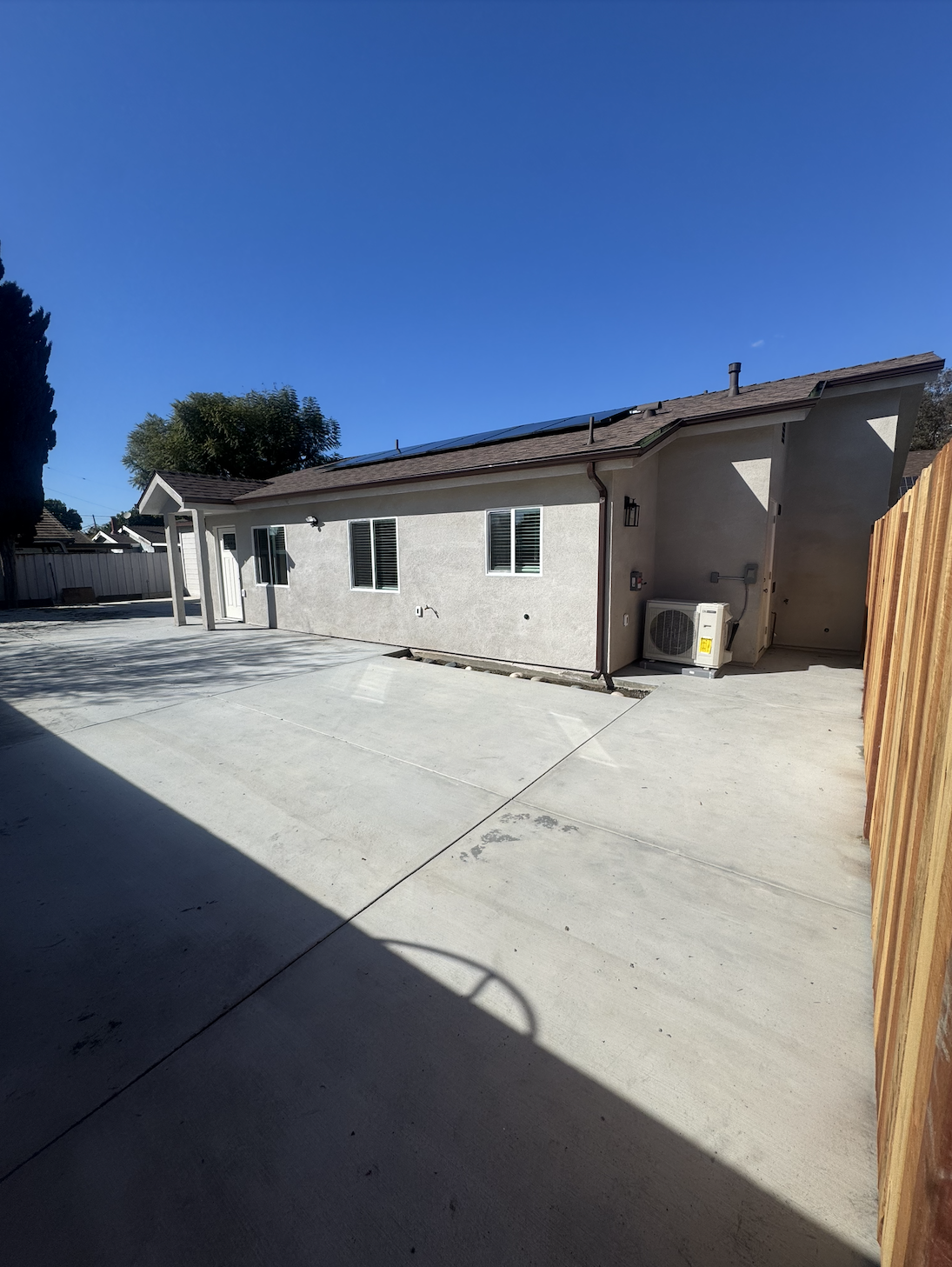 Backyard with concrete patio, house with white walls, three windows, door, solar panels on roof, air conditioning unit, and wooden fence on the right under clear blue sky.