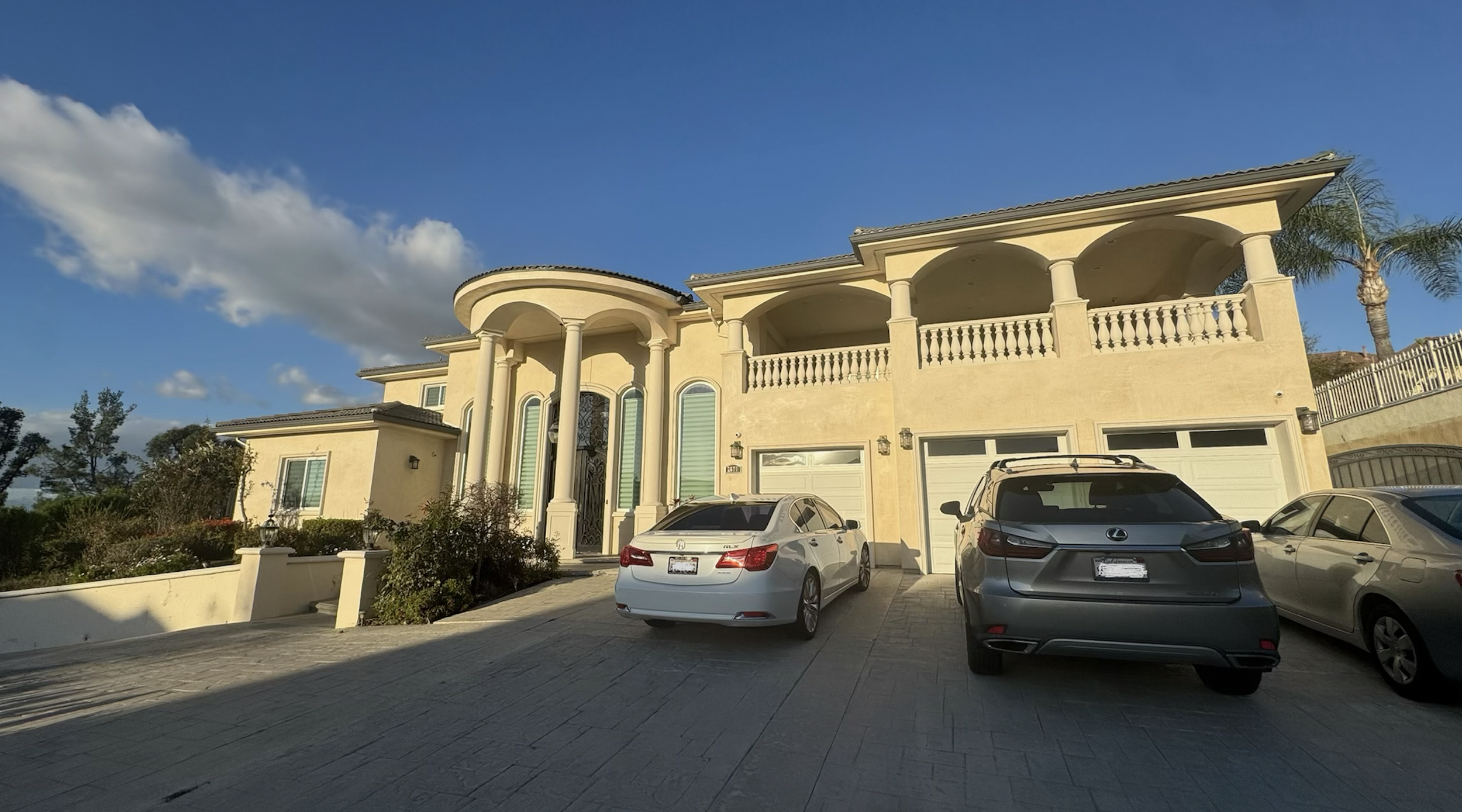 Large, elegant house with columns, balconies, and a beige facade, with three parked cars in the driveway under a blue sky with clouds.