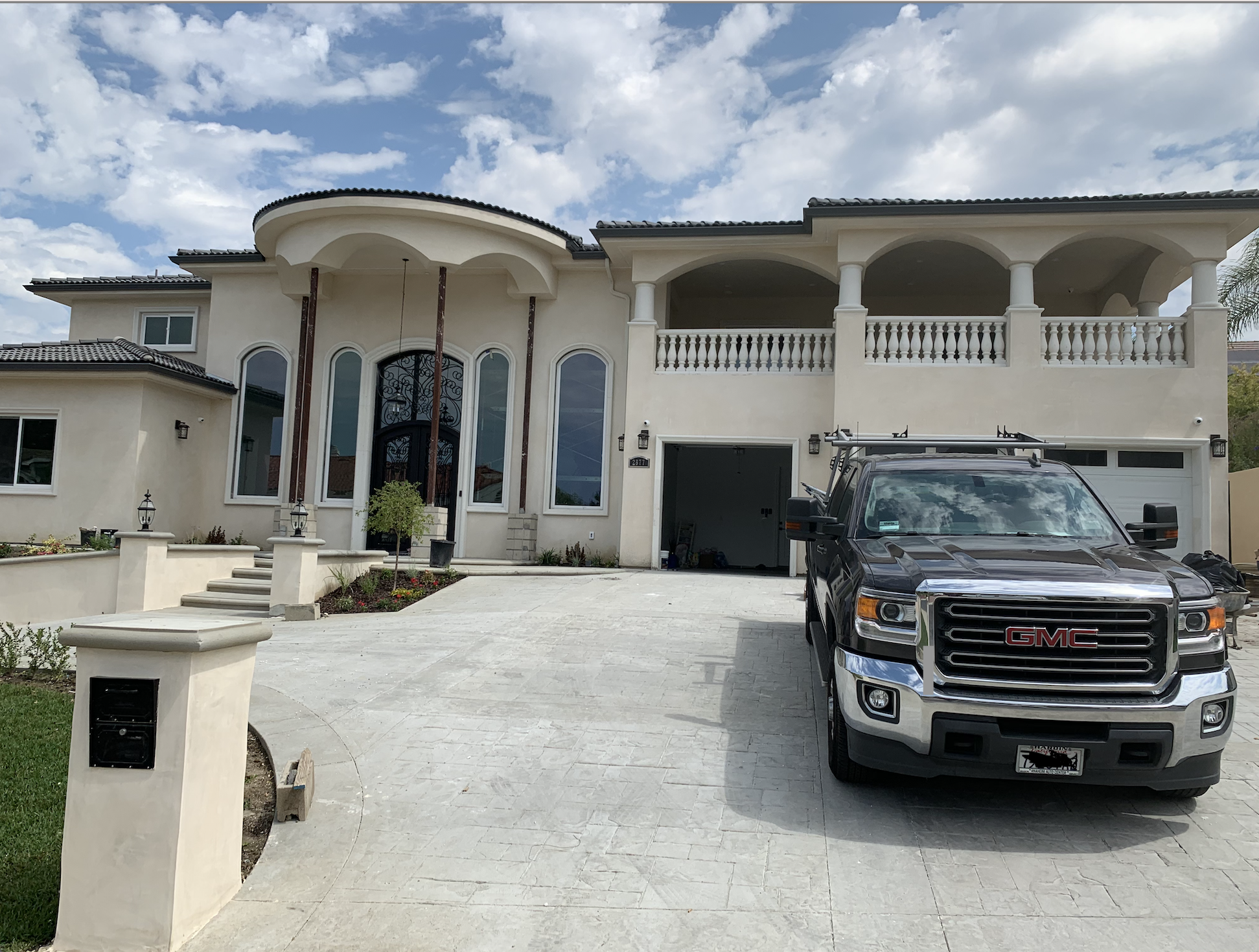 Front view of a large, modern two-story house with a driveway, a GMC truck parked in front, and a garage. The house features tall windows, a balcony with balustrades, and a small tree near steps leading to the front door.