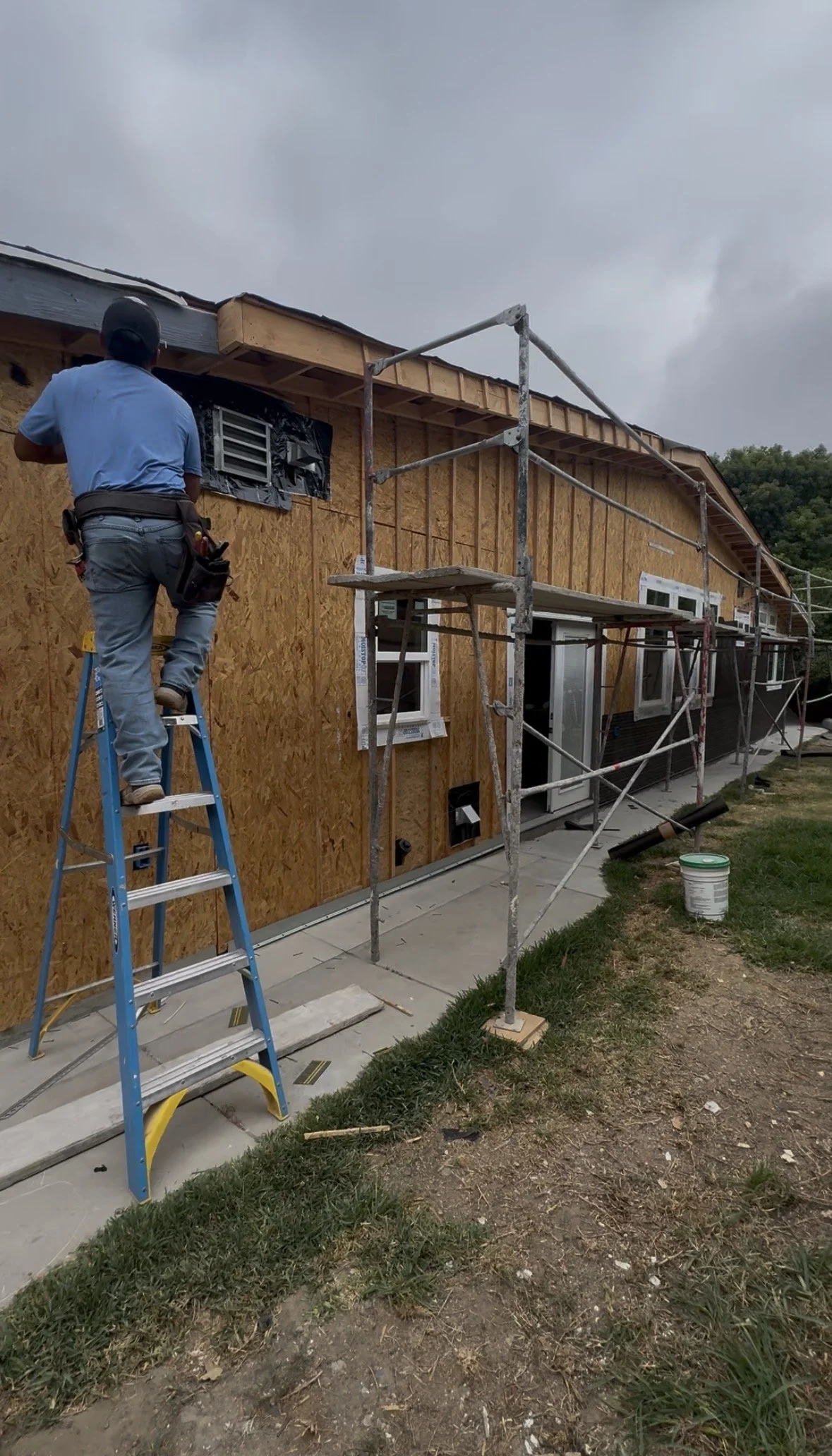 Construction worker standing on a ladder installing or inspecting the exterior wall of a house, which is covered with wooden sheathing and electrical wiring, with scaffolding set up along the side of the house.