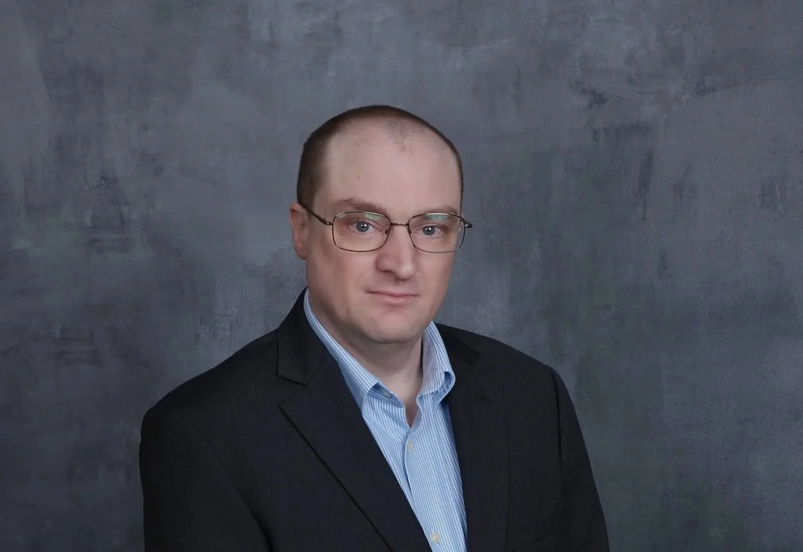 A professional headshot of a man wearing glasses, a dark blazer, and a light blue collared shirt, with a textured gray background.