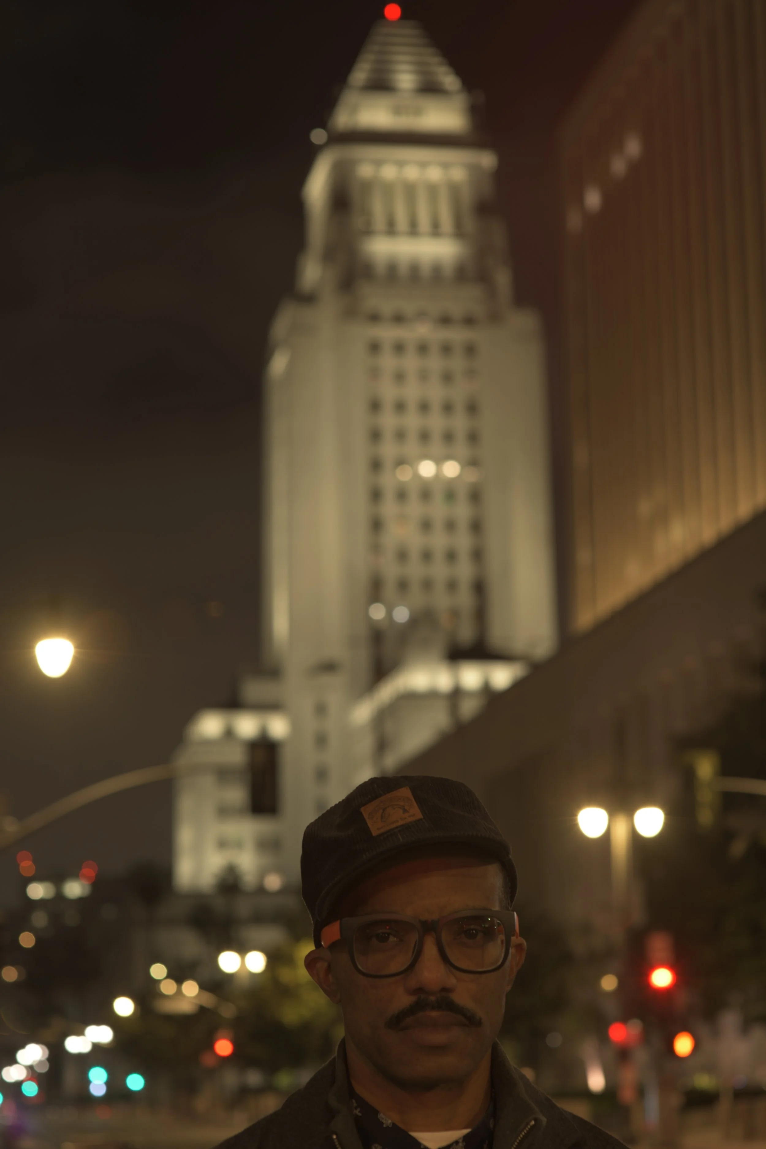 A nighttime city scene featuring a man with glasses and a cap in the foreground, with a tall, illuminated skyscraper in the background.