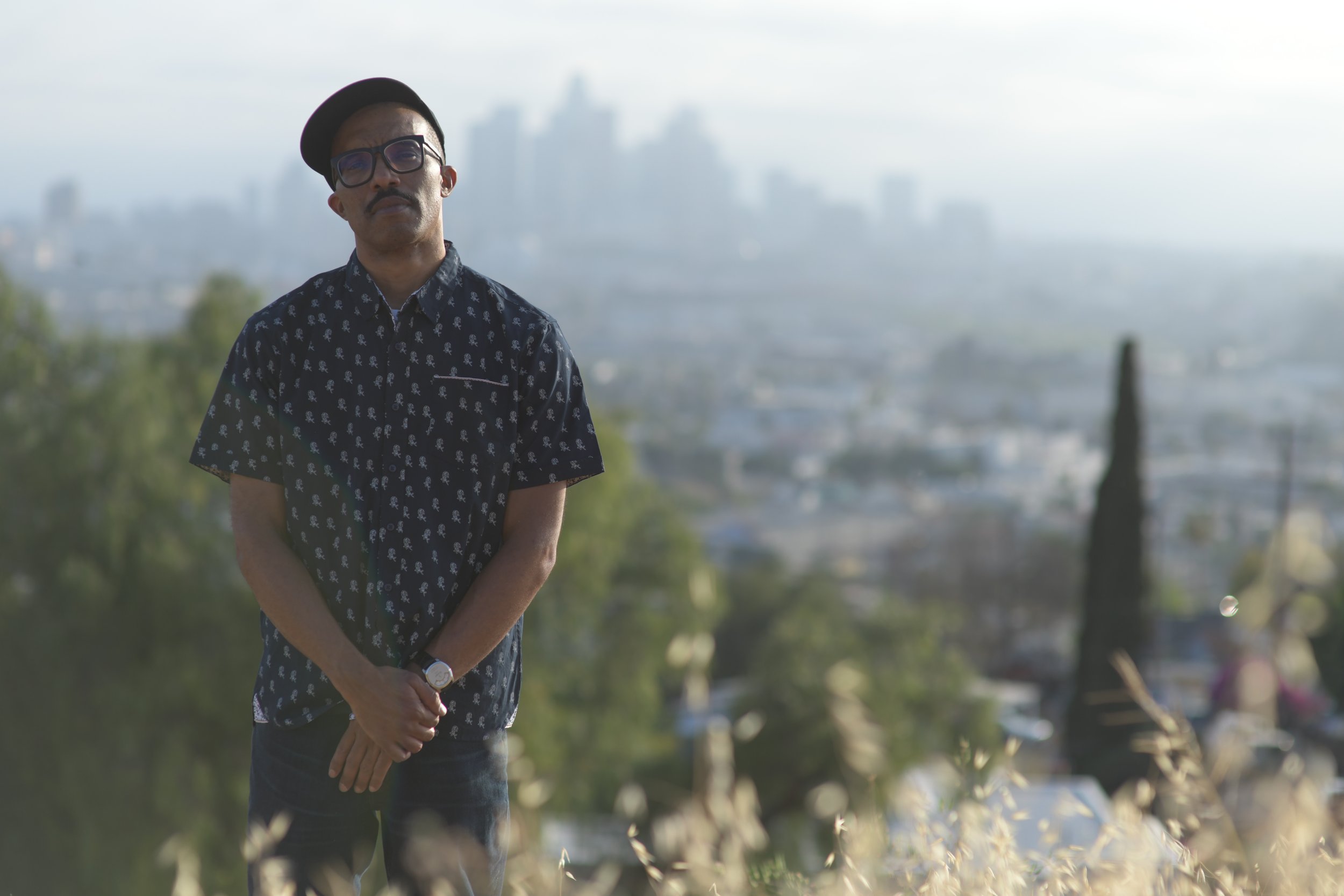 A man standing outdoors on a hilltop, wearing glasses, a patterned short-sleeve shirt, a watch, and a beret, with blurred cityscape in the background.