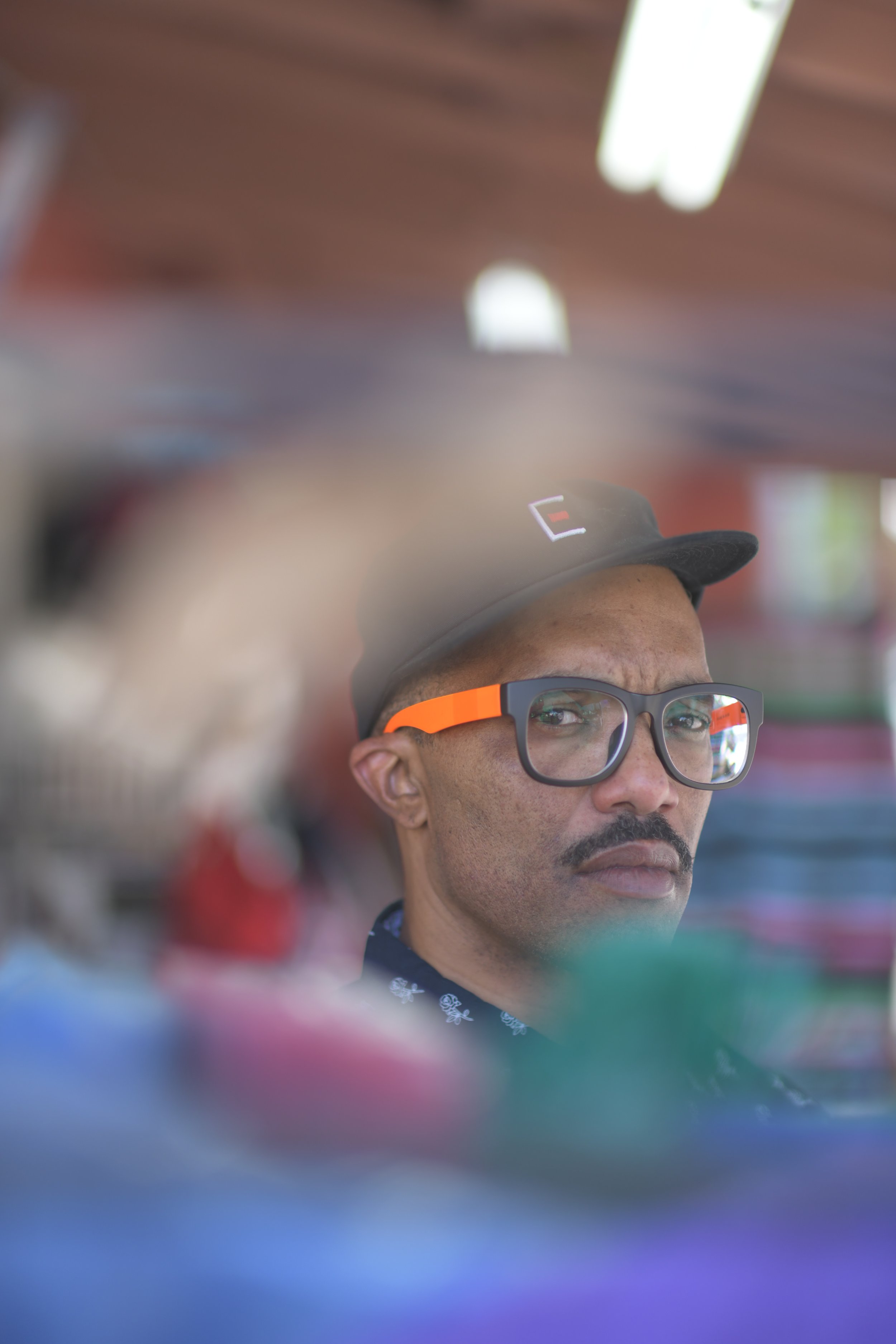 A man with glasses and a black cap looking at the camera, reflected in a mirror or a glass surface with blurred colorful objects in the foreground.
