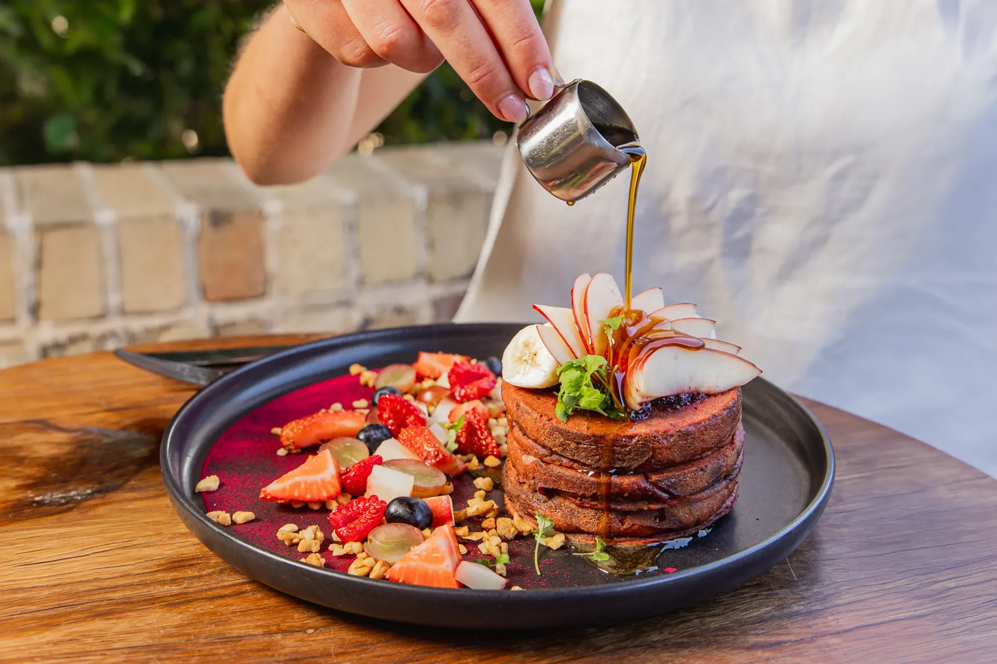 A stack of chocolate pancakes topped with sliced bananas, apples, and fresh herbs, with syrup being poured over. A side of mixed berries and chopped nuts on a black plate on a wooden table.