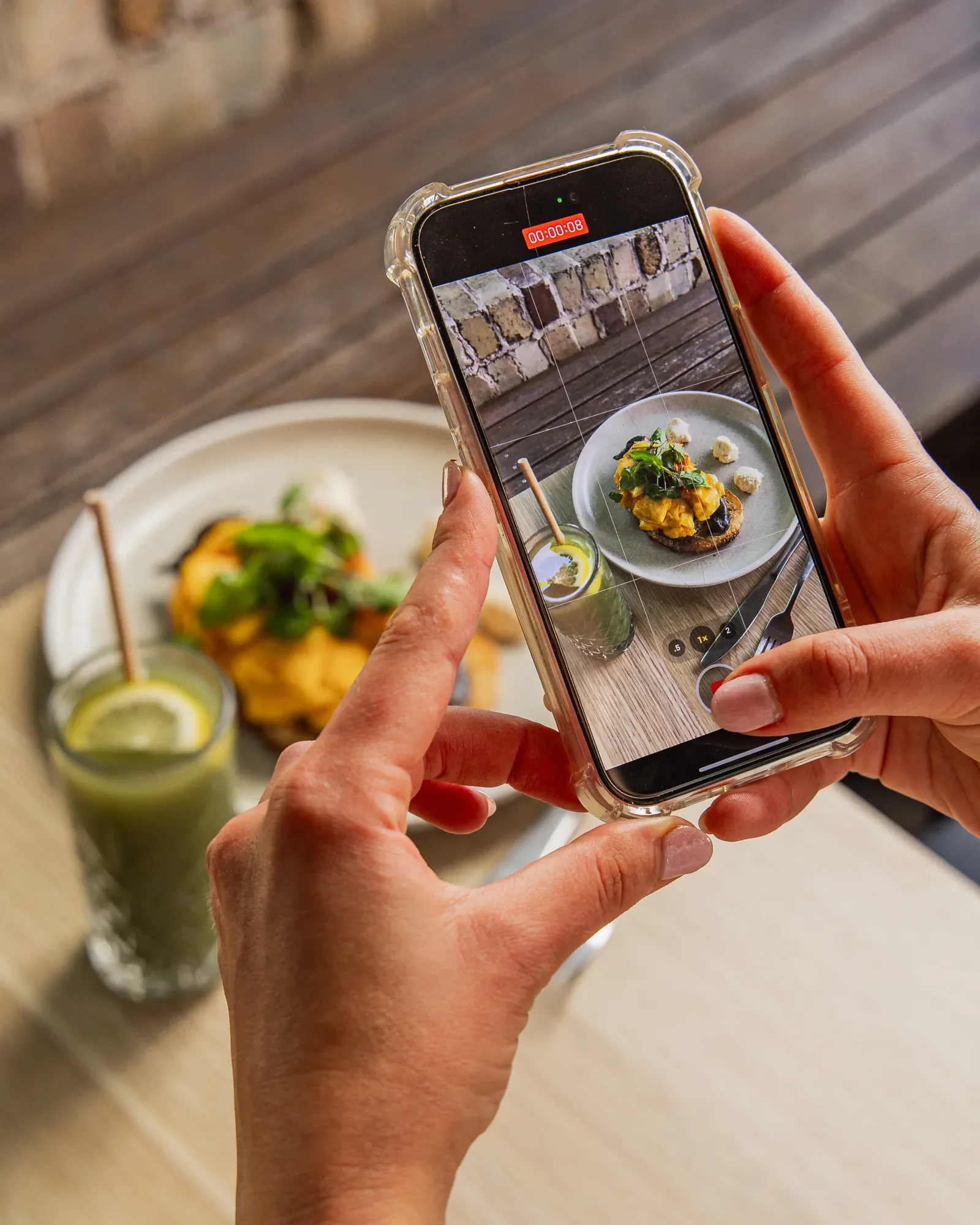 Person taking a photo of a plate of food and drinks on a wooden table with a smartphone.