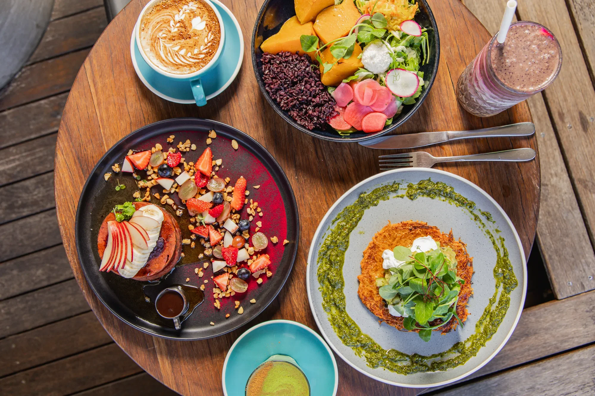 A wooden table with various dishes: a fruit tart with sliced strawberries, bananas, blueberries, and nuts; a salad with mixed greens, radish slices, and melon chunks; a plate with shredded meat topped with microgreens and a green sauce; a bowl of smo