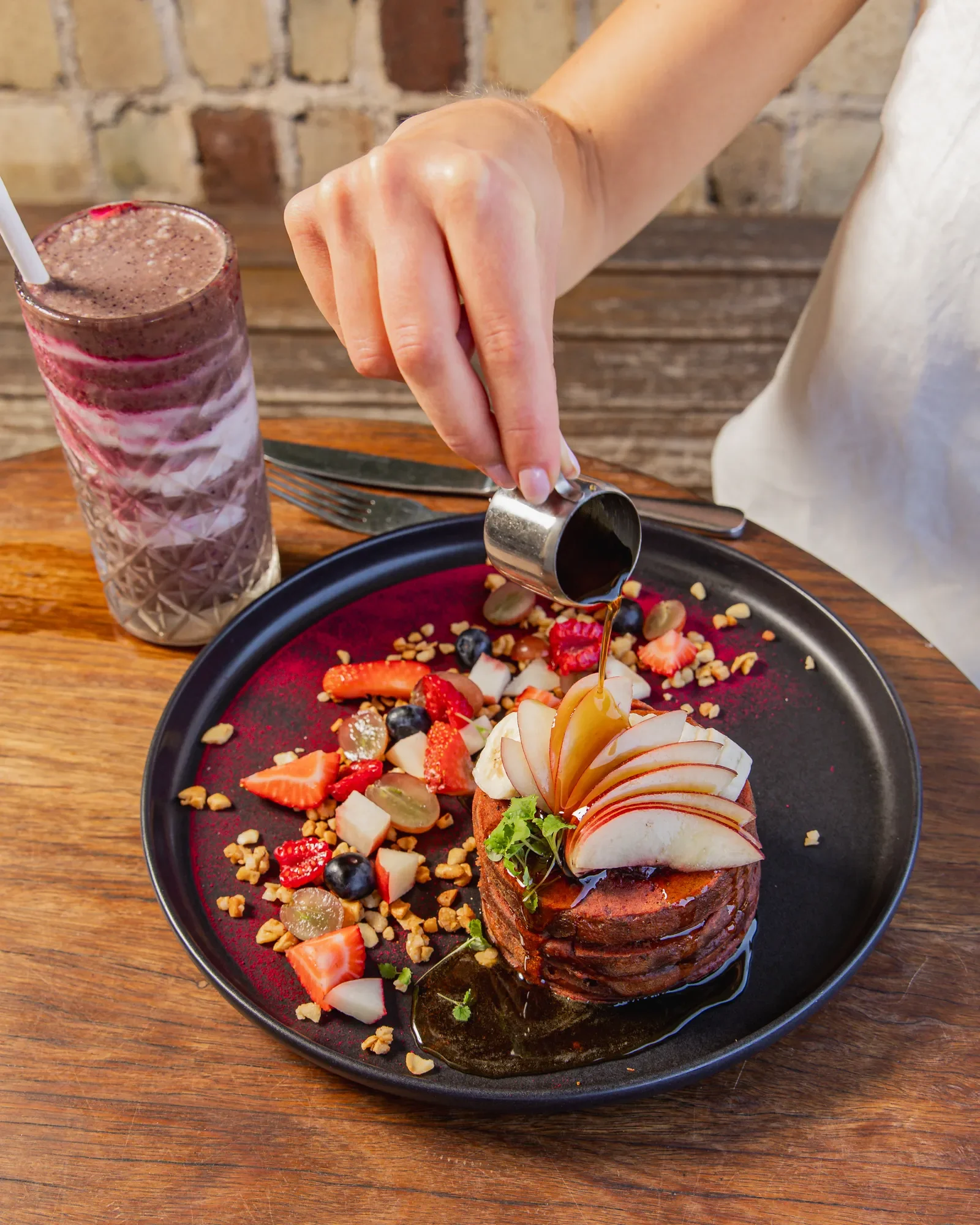 Person pouring syrup over a stack of pancakes topped with sliced apples and garnished with greenery, accompanied by mixed fruit and crunchy toppings, with a berry smoothie on the side.