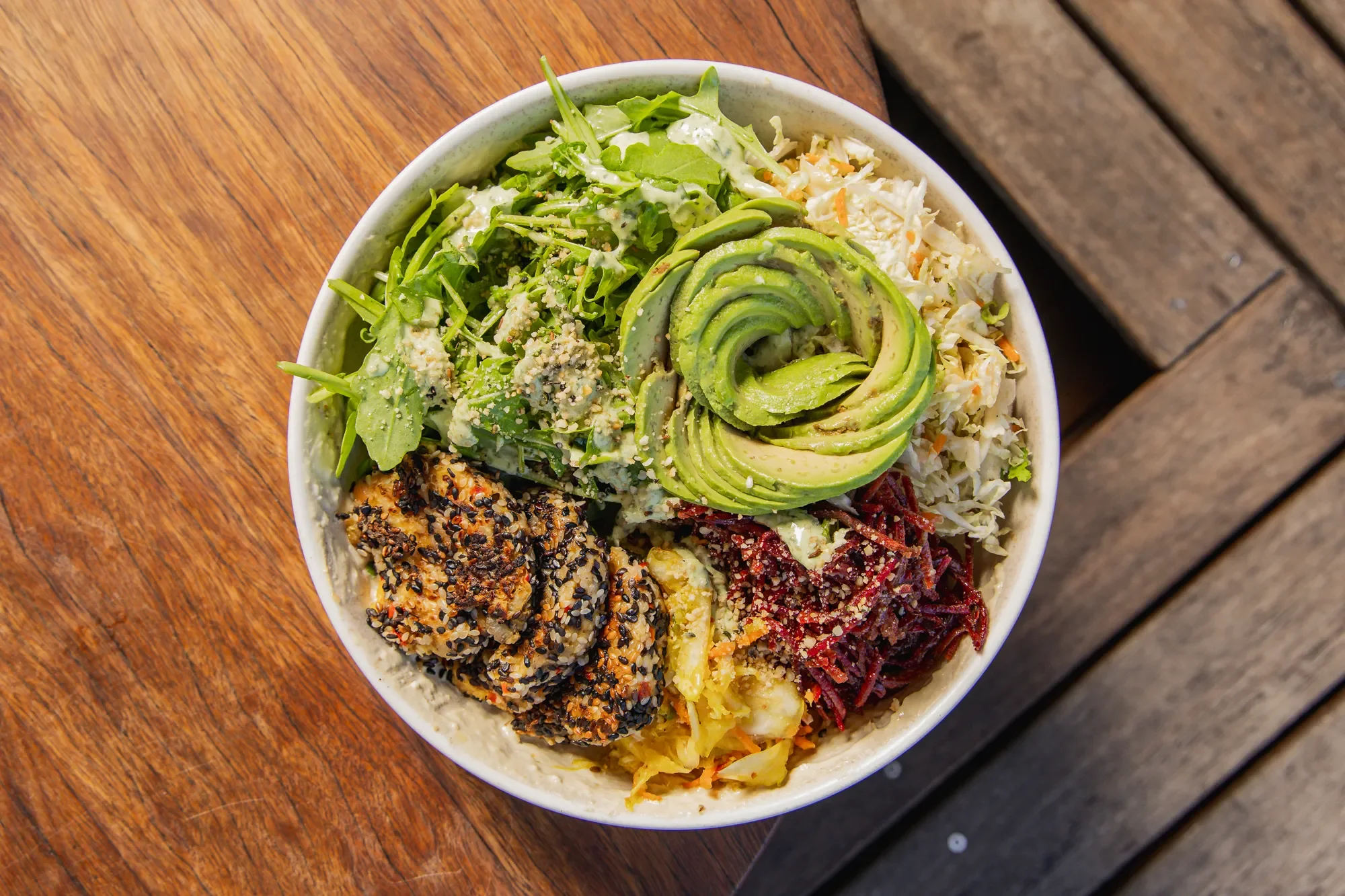 A bowl filled with various salads, including sliced avocado, greens, shredded cabbage, and a sesame-crusted protein, placed on a wooden table.