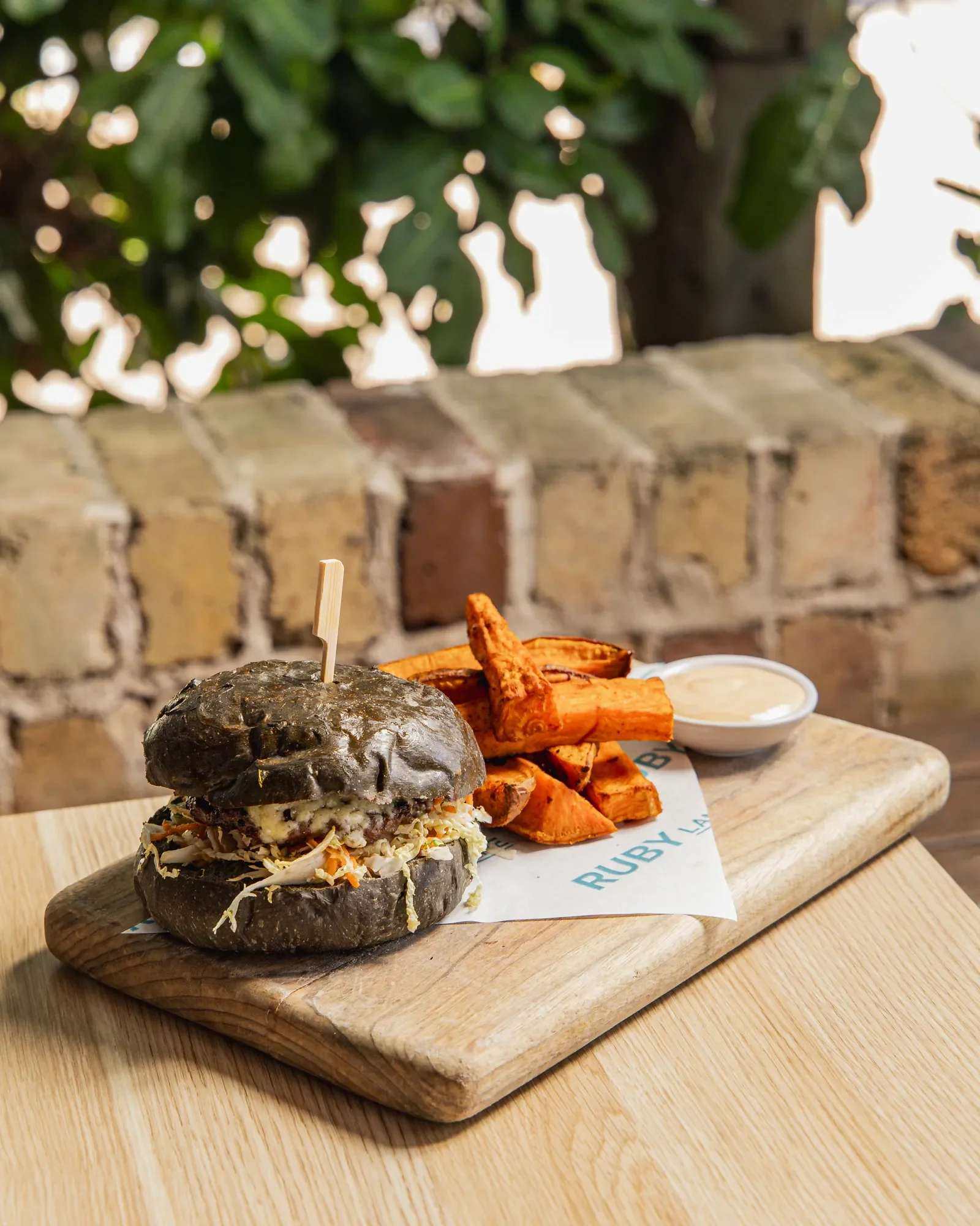 A burger with black bun, lettuce, and sauce on a wooden board, served with sweet potato fries and a small bowl of dipping sauce at Ruby's