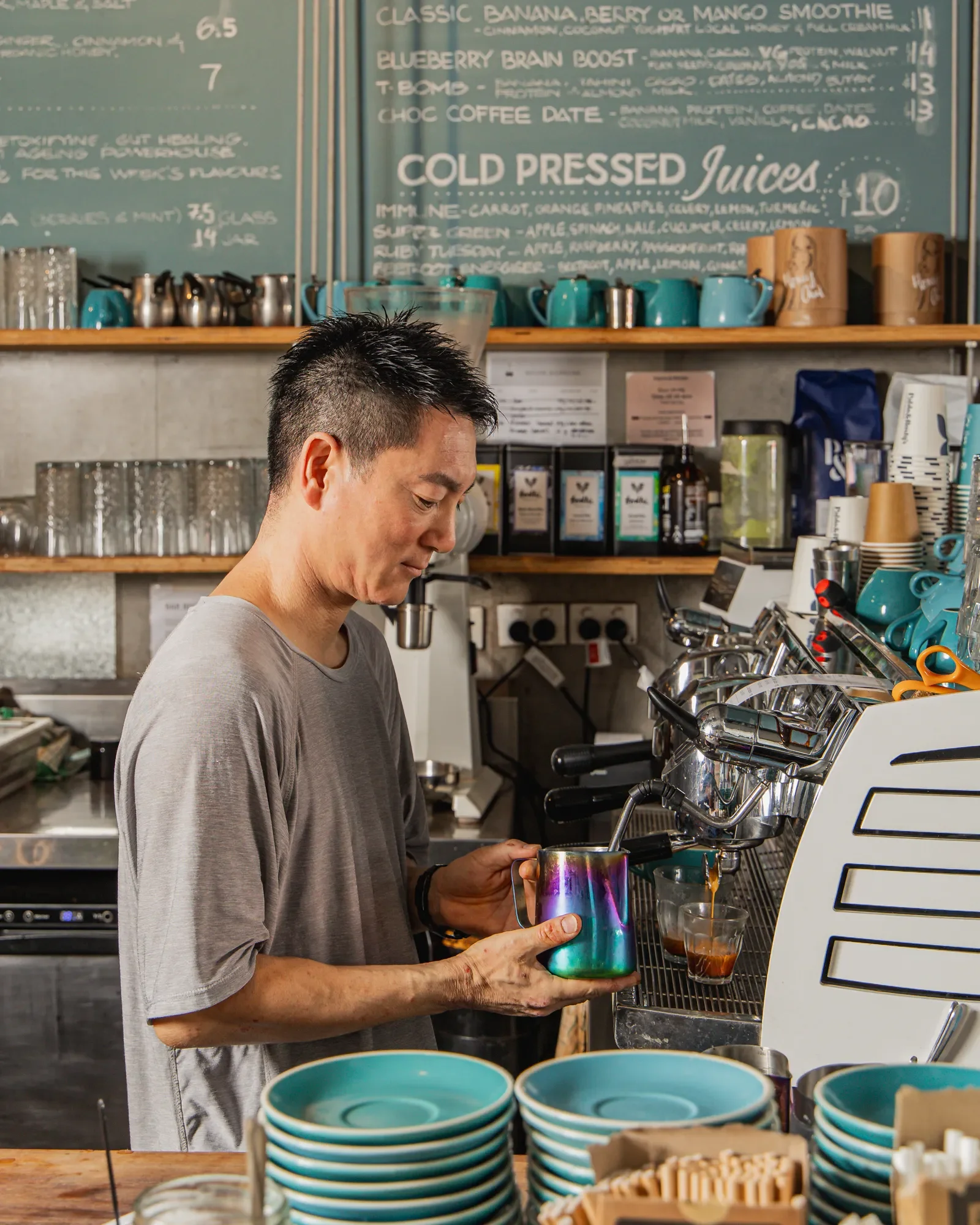A barista pours coffee from an espresso machine into a metallic cup, surrounded by stacks of teal bowls and various coffee-making supplies in a coffee shop.
