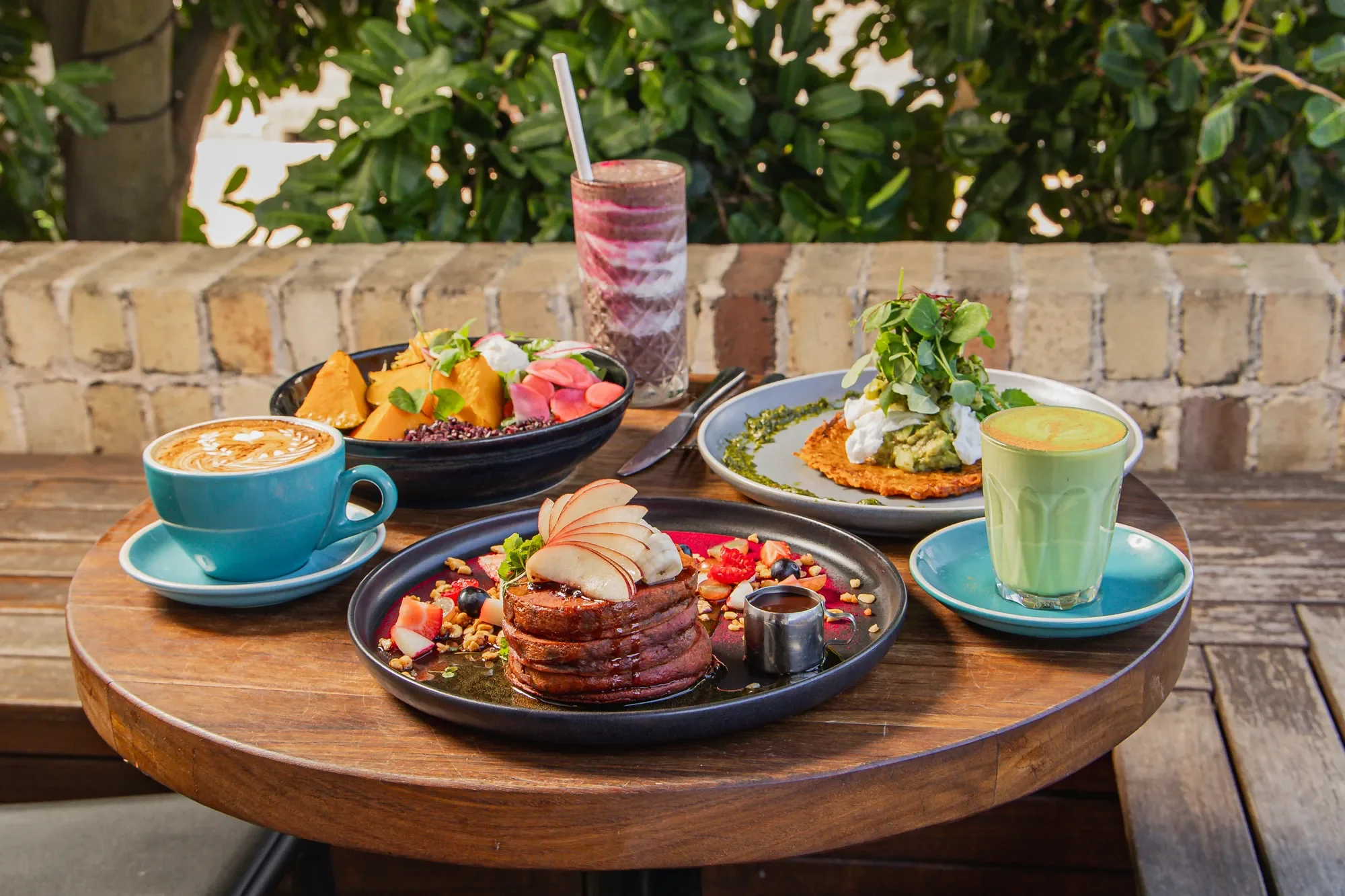 Assorted breakfast foods on a wooden table including a cup of coffee, a tall glass of berry smoothie, a green-colored beverage, a bowl of fruit and yogurt, a plate of pancakes with fruit and syrup, and a savory dish with sauce and greens, set outdoor