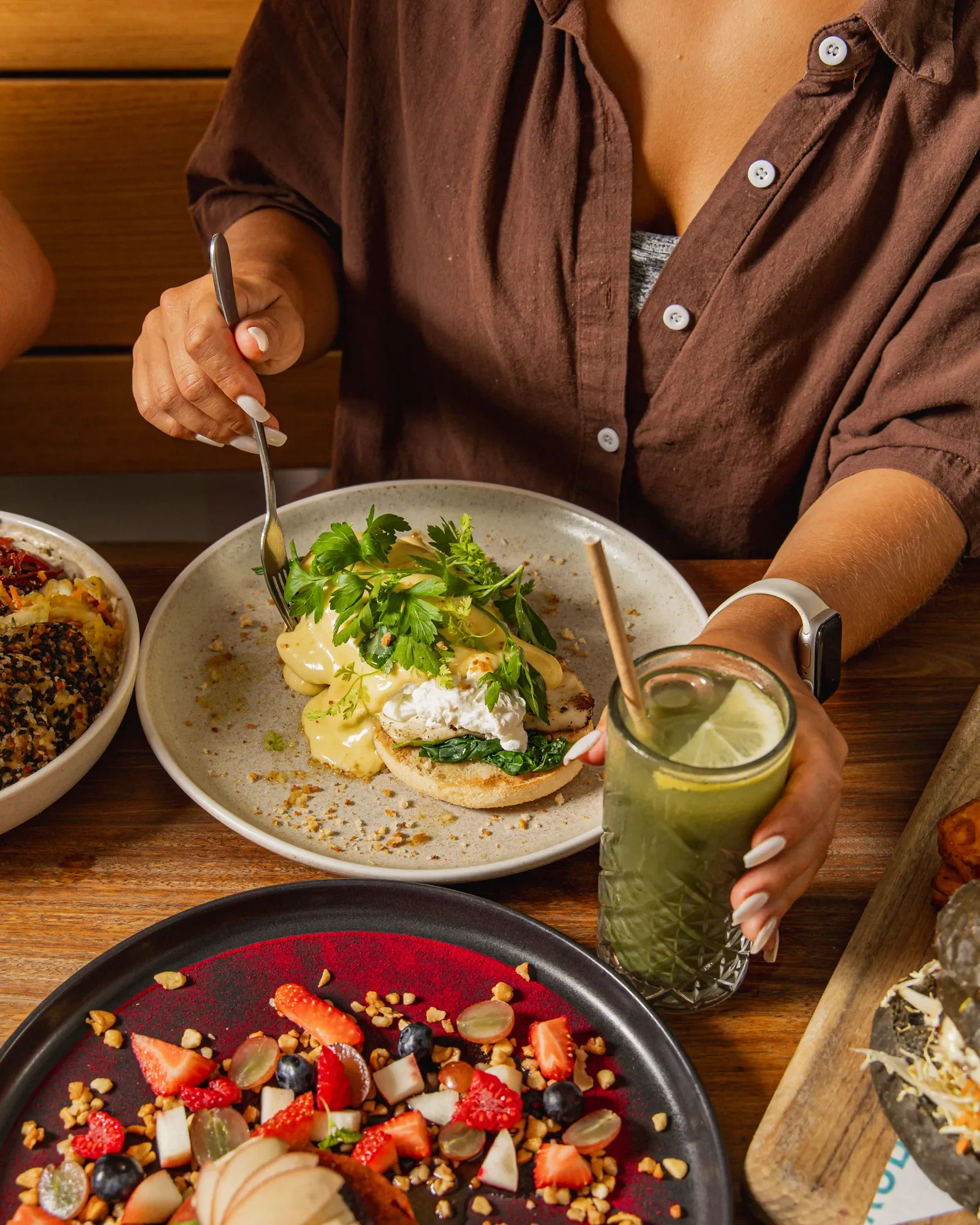 A person is holding a fork and a drink, sitting at a table with various foods including a bagel with toppings, a salad, and a fruit platter.