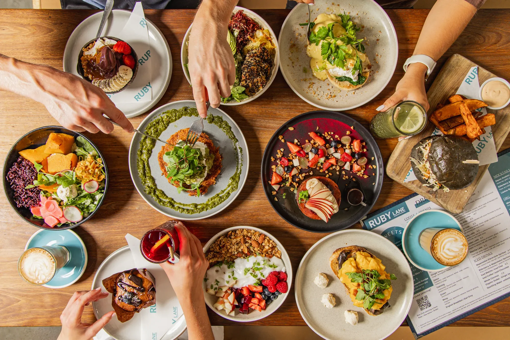 A variety of colorful desserts and dishes on a wooden table, including salads, fruit, cake, and drinks, with hands reaching for food.